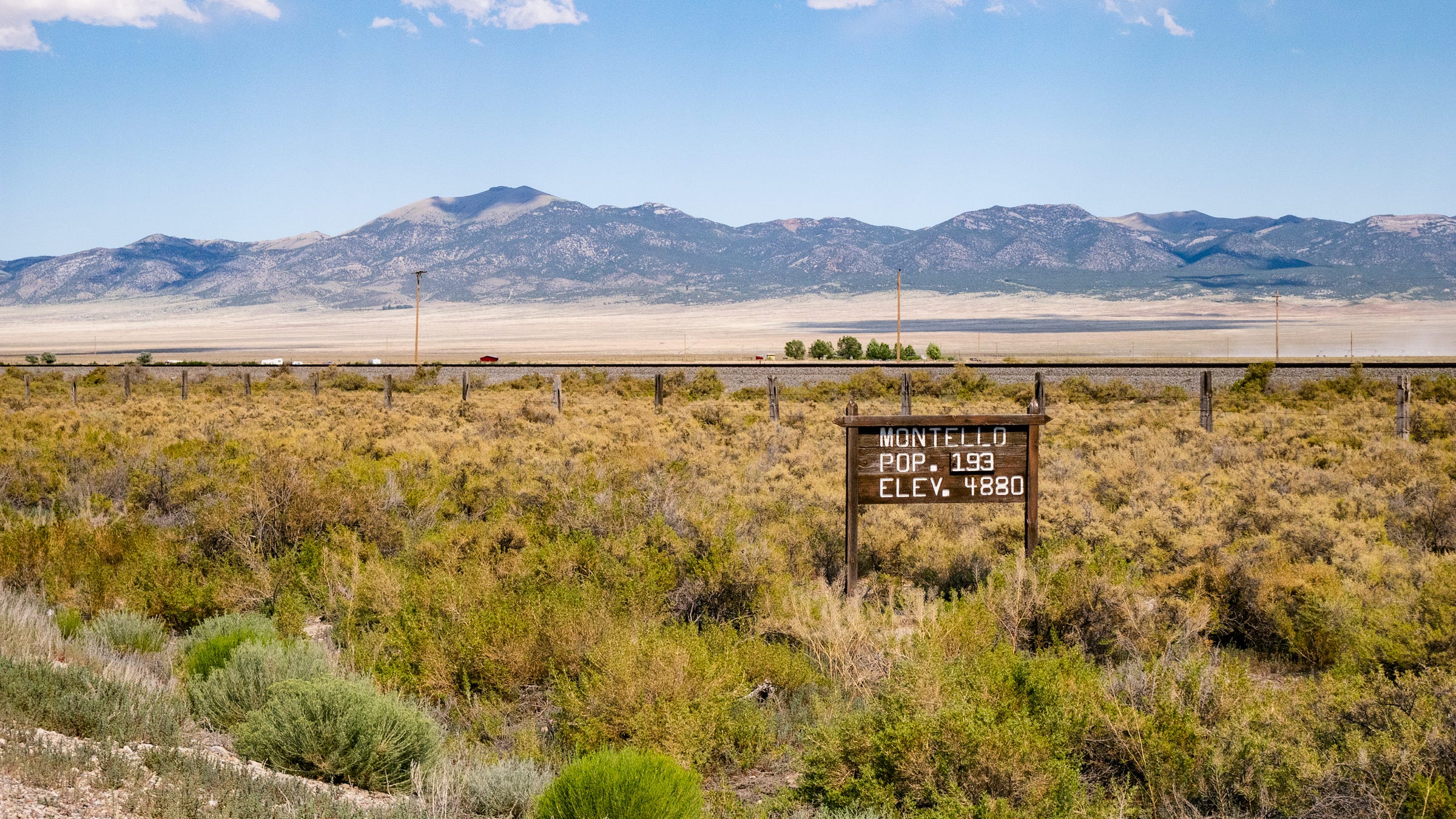 Photo of sage and other low brush in the foreground, the railroad tracks, a broad valley, and dark jagged mountains in the distance. In the foreground is a simple wooden sign, "MONTELLO / POP. 193 / ELEV. 4880