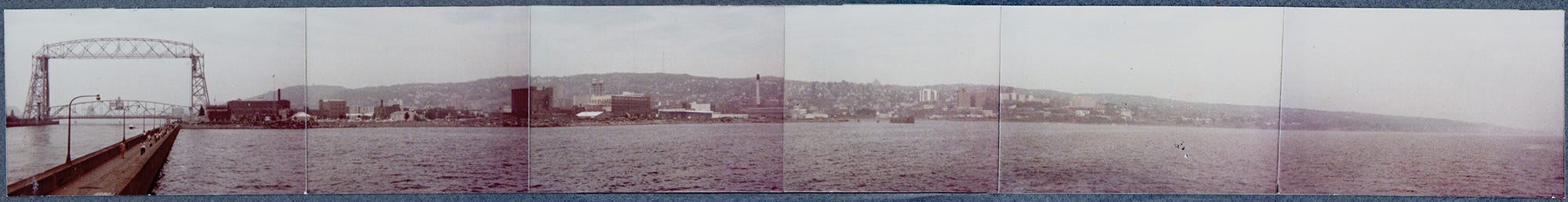 A six-frame panorama of Duluth, Minnesota taken in August of 1978 with a 110-film camera from the North Pier Lighthouse. The six frames are mounted on blue matt board, and many of the prints show signs of wear from several moves and improper storage. At the far left of the frame is the Aerial Lift Bridge, with the city taking up most of the next four frames.