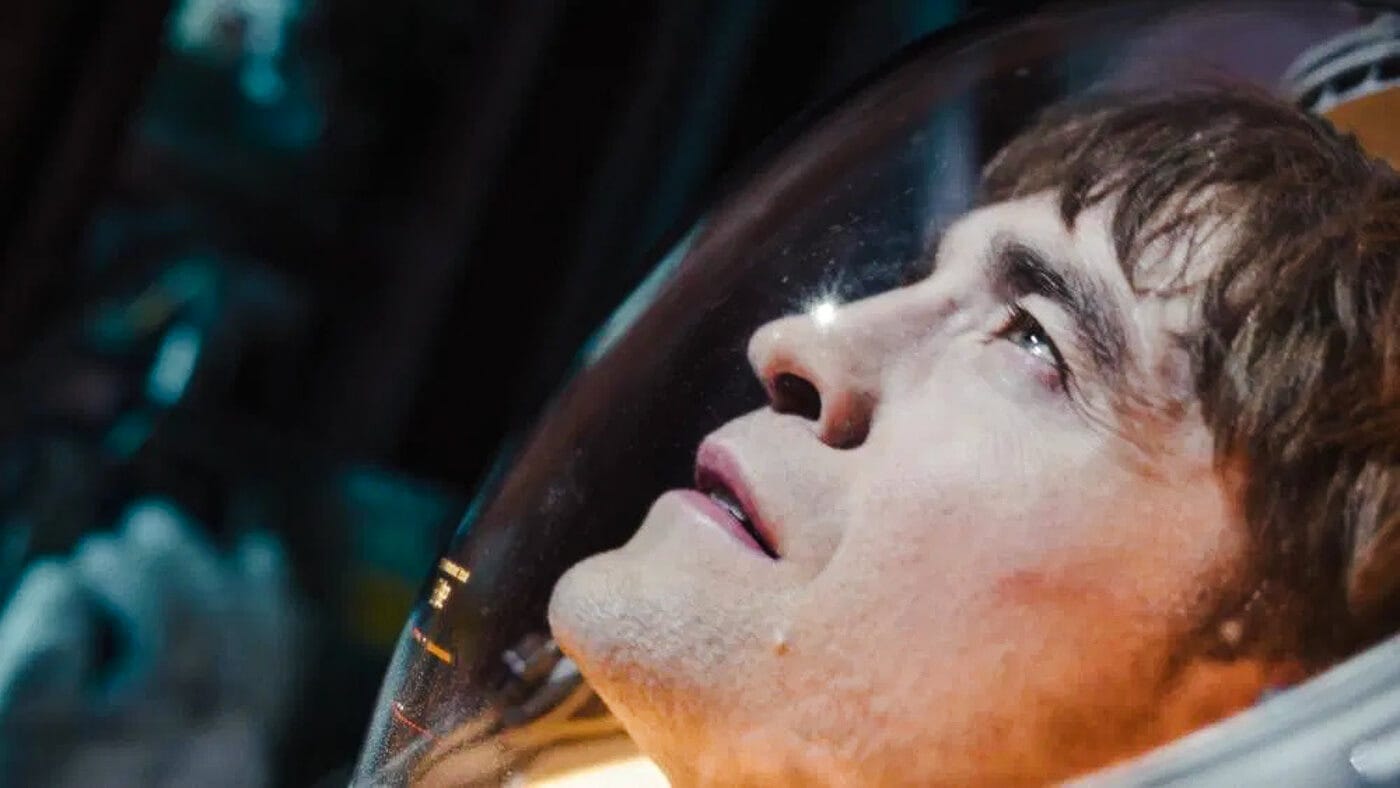 Close-up of Robert Pattinson’s face in a space helmet, staring upward with a mix of resignation and quiet dread as cold light reflects off the visor.