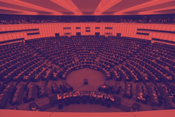 The inside of the European Parliament. There is semicircle of raked seating filled with parliamentarians and a red filter over the image.