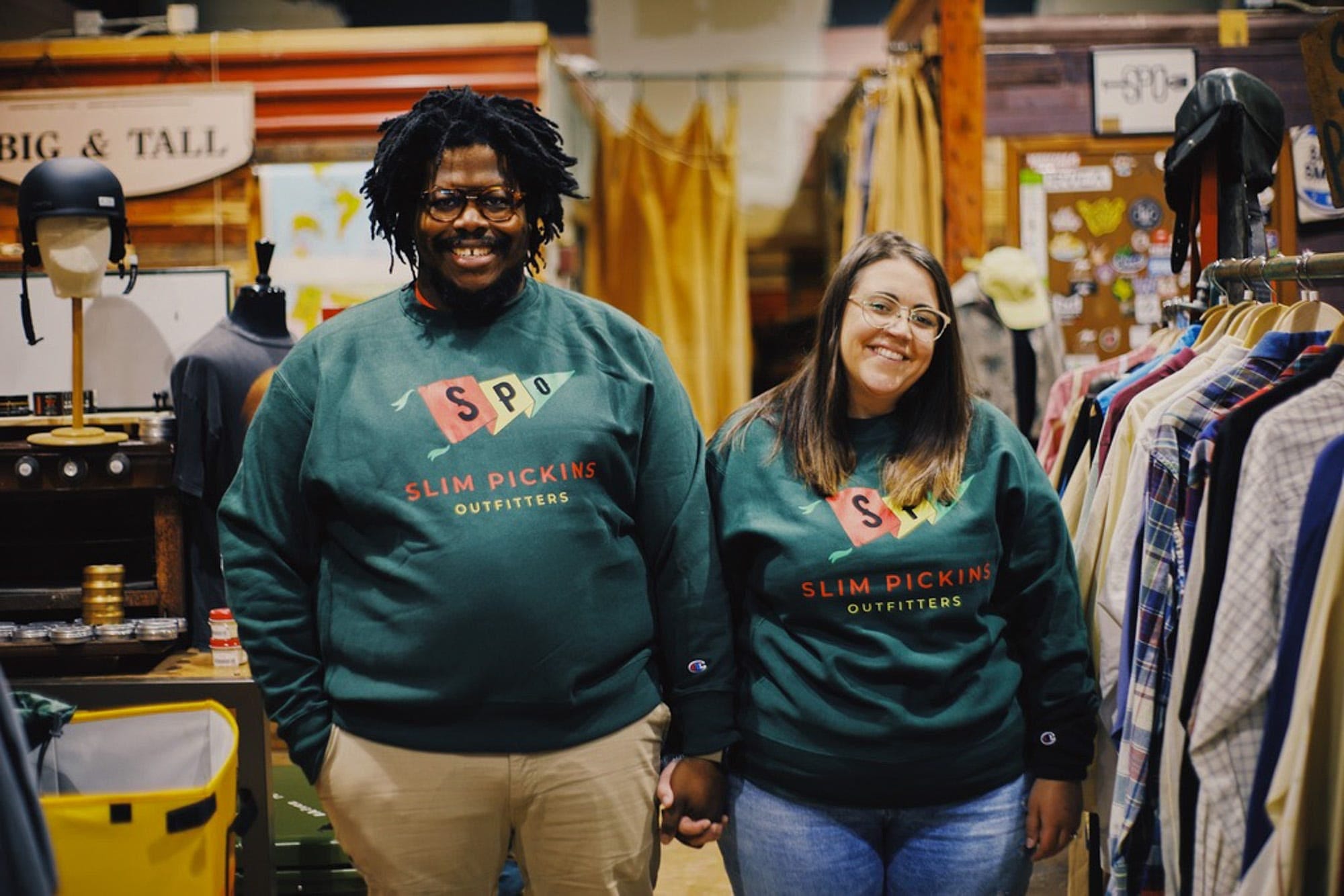 Two figures in matching green Slim Pickins Outfitters sweatshirts, one a Black man with medium-length hair and glasses and the other a white woman with brown hair and glasses, hold hands and face the camera in a three-quarter-length portrait taken inside an outdoor-gear store