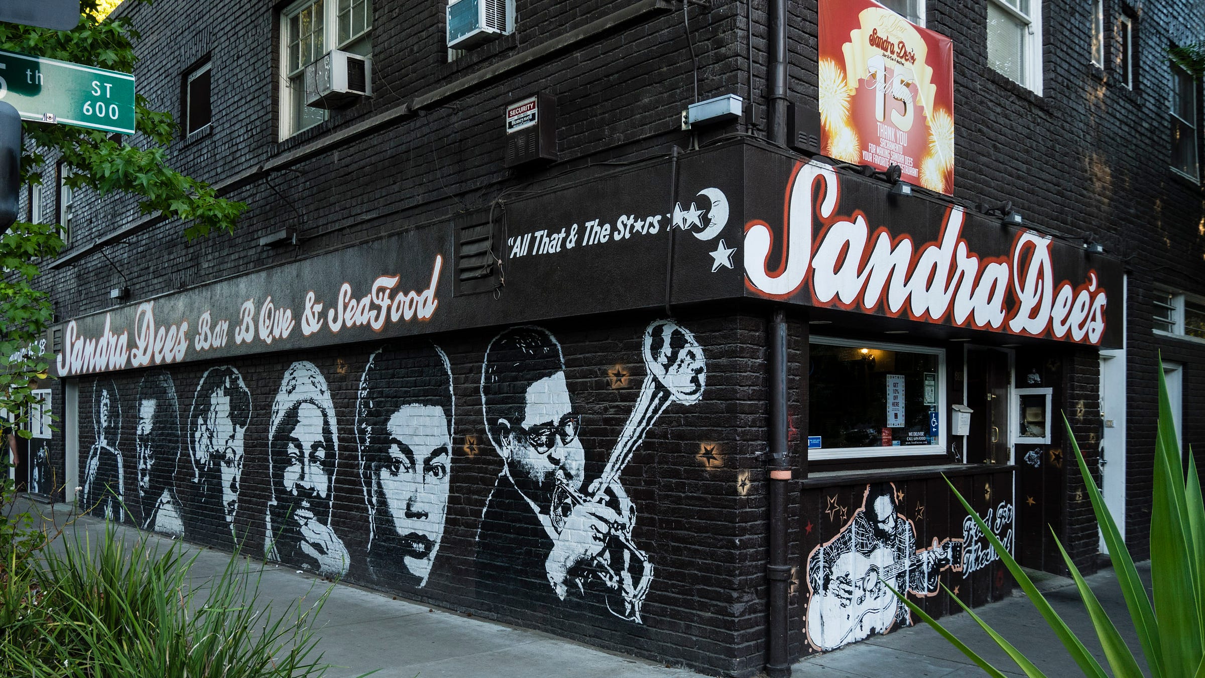 Sandra Dee's Bar B Que and Sea Food (written in thick, stylized script, white with an orange outline) is on the corner of a brown brick building. The long wall is illustrated with oversized portraits of jazz giants including Miles Davis, Billie Holiday, Nina Simone (I think) Dizzy Gillespie, and others. 