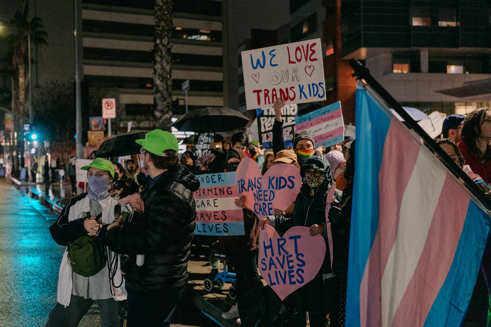 Protestors outside of CHLA hold signs and trans flags. Signs say "WE LOVE OUR TRANS KIDS", "TRANS KIDS NEED CARE" "HRT SAVES LIVES" and similar.