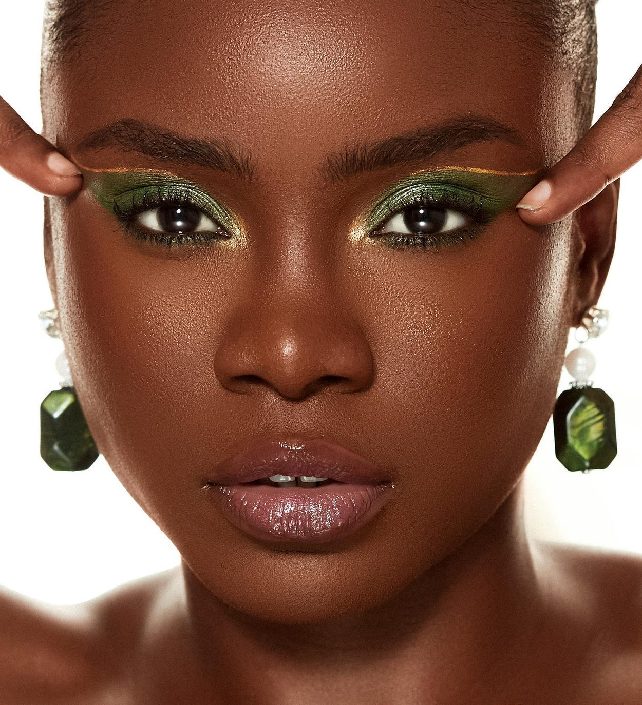 A woman of African descent poses with one finger pressed to her eye linein a front-on headshot. She’s wearing green eyeshadow with a gold streak, and decorative white, black, and green earrings. The backdrop is white. A woman of African descent poses with one finger pressed to her eye linein a front-on headshot. She’s wearing green eyeshadow with a gold streak, and decorative white, black, and green earrings. The backdrop is white.
