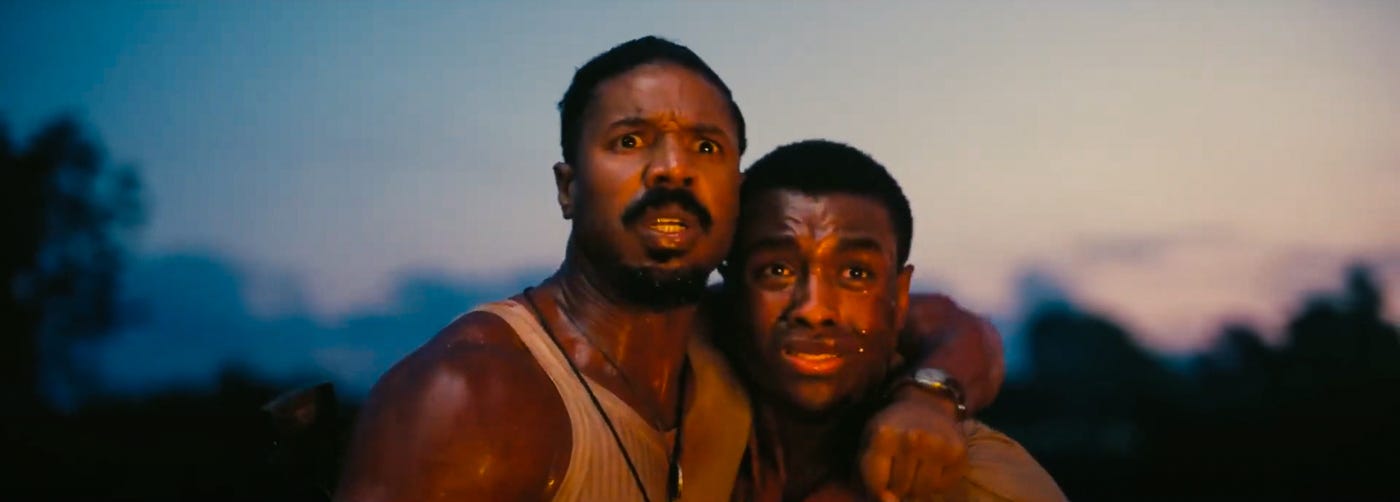 Michael B. Jordan and Miles Caton stand locked in an embrace against a dusky sky, both glowing and glistening with sweat and staring ahead in horror in a harrowing moment.