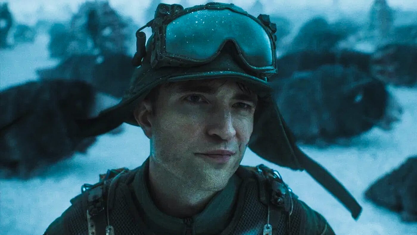 Robert Pattinson stands in snowy terrain wearing tactical gear and frost-covered goggles, smirking slightly despite the bleak, alien battlefield around him.
