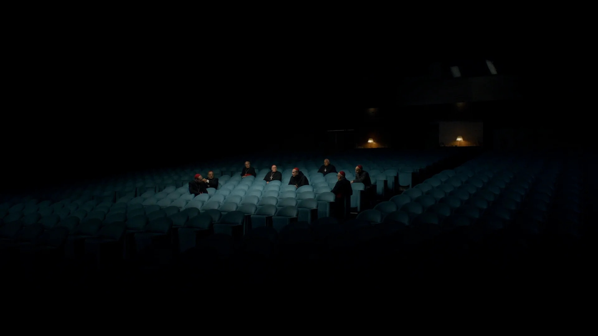 a group of cardinals sitting in a halo of chairs in a dark auditorium in Conclave