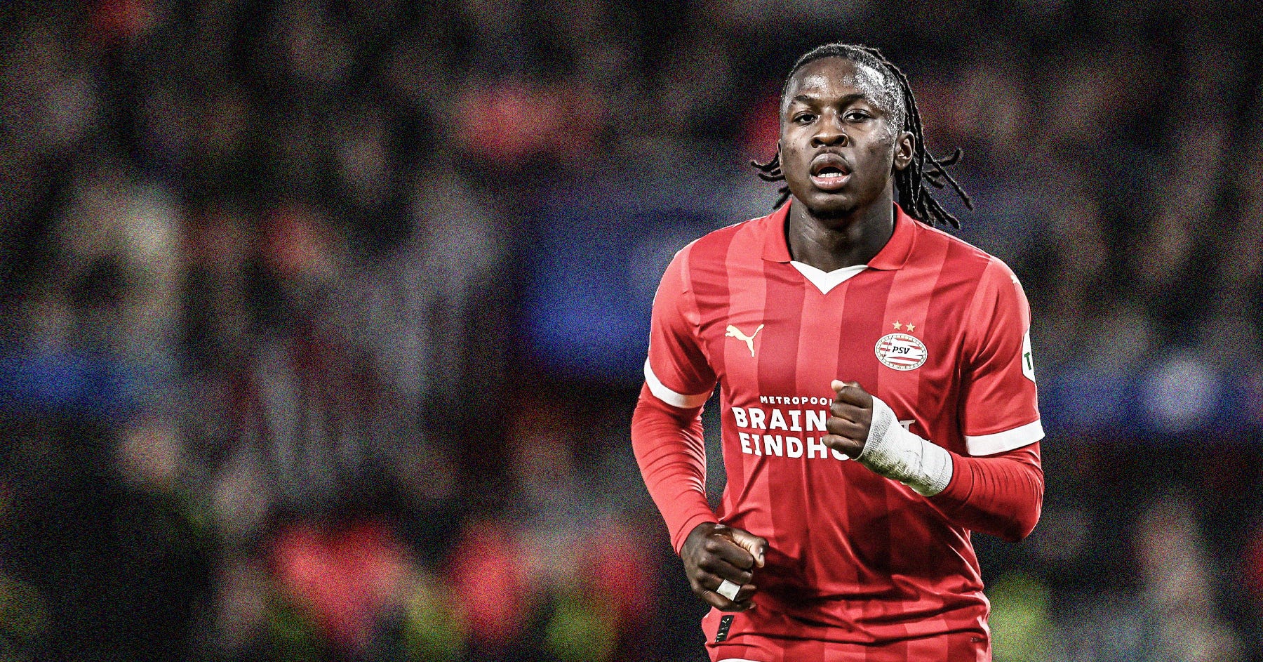 A photo of Johan Bakayoko running in a red PSV Eindhoven shirt set against a blurred crowd background.