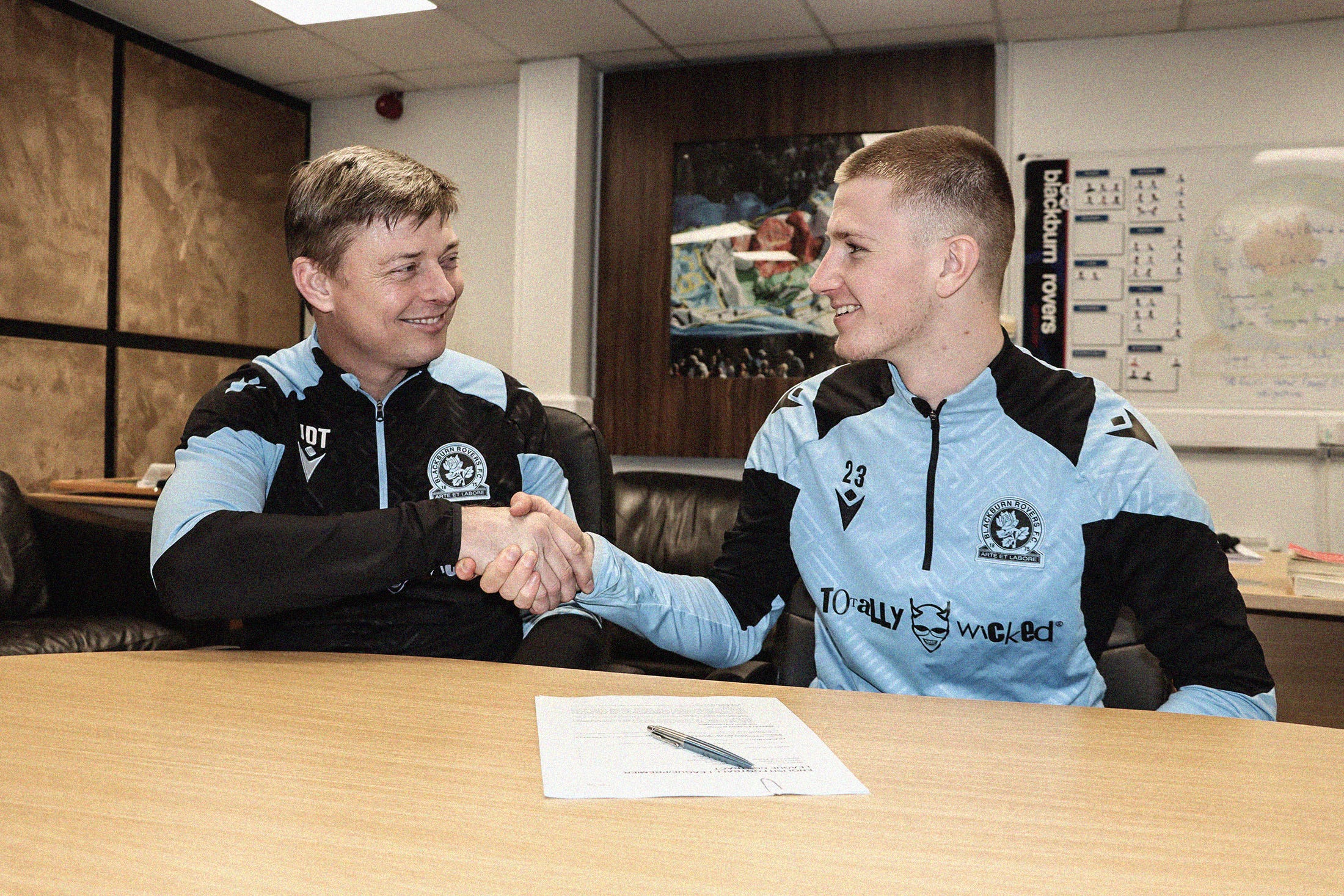 A photo of Jon Dahl Tomasson and Adam Wharton, shaking hands, looking and smiling at each other while sat at a table, with a signed contract in the foreground and an office scene in the background.