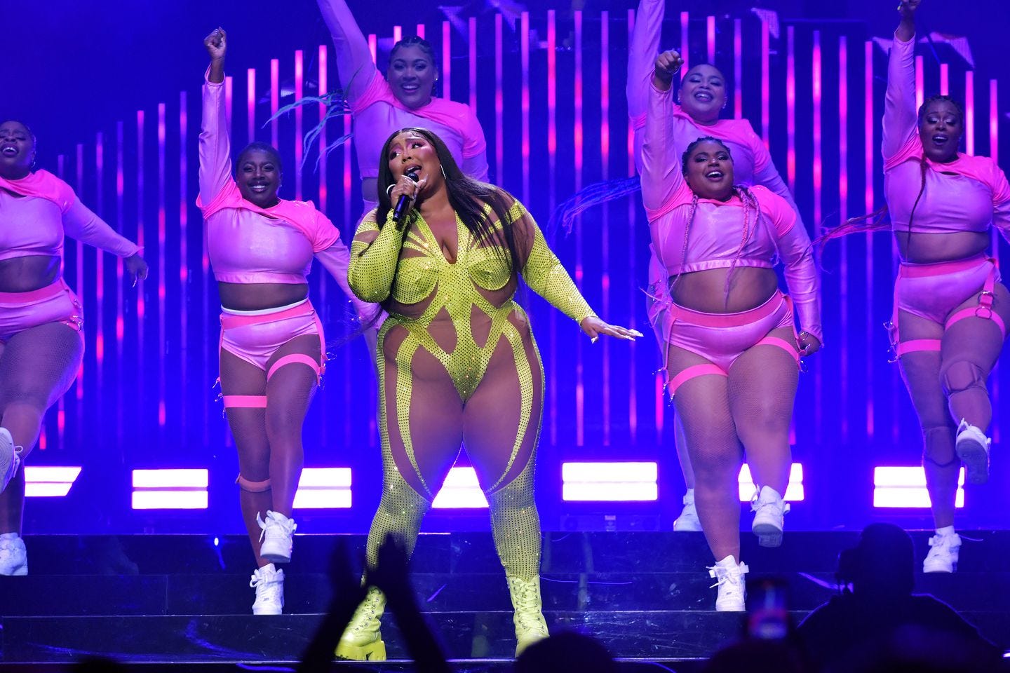 Lizzo performs onstage in a sparky yellow costume, with her dancers behind her in pink athletic costumes