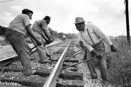Gandy dancers (railroad workers), Mississippi, 1976  Roland L. Freeman  The Museum of Fine Arts, Houston  Black and white photo of men using long steel bars to reposition the rails of a railroad track. Three of the men are alongside the near rail, and another three alongside the far rail. Three of the men closest to the camera are visible and recognizable as African American. The other three are obscured by the three men in front but their long steel bars can ben seen. The men are wearing worn and dirty clothes - slacks, shirts, and hats, but the man closest to the camera has finer trousers, a long-sleeved collared shirt, suspenders, and a straw porkpie hat. All six men have placed one end of the steel bar beneath the iron rail, and with a firm grip on the other end of the bar are straining their backs to reposition the rail. There is gravel ballast at their feet and tall wild grasses along the edge of the tracks. In the distance, supervising the work, is a white man with dark slacks and a white short sleeve shirt. He is standing with his feet shoulder width apart and watching the six men.