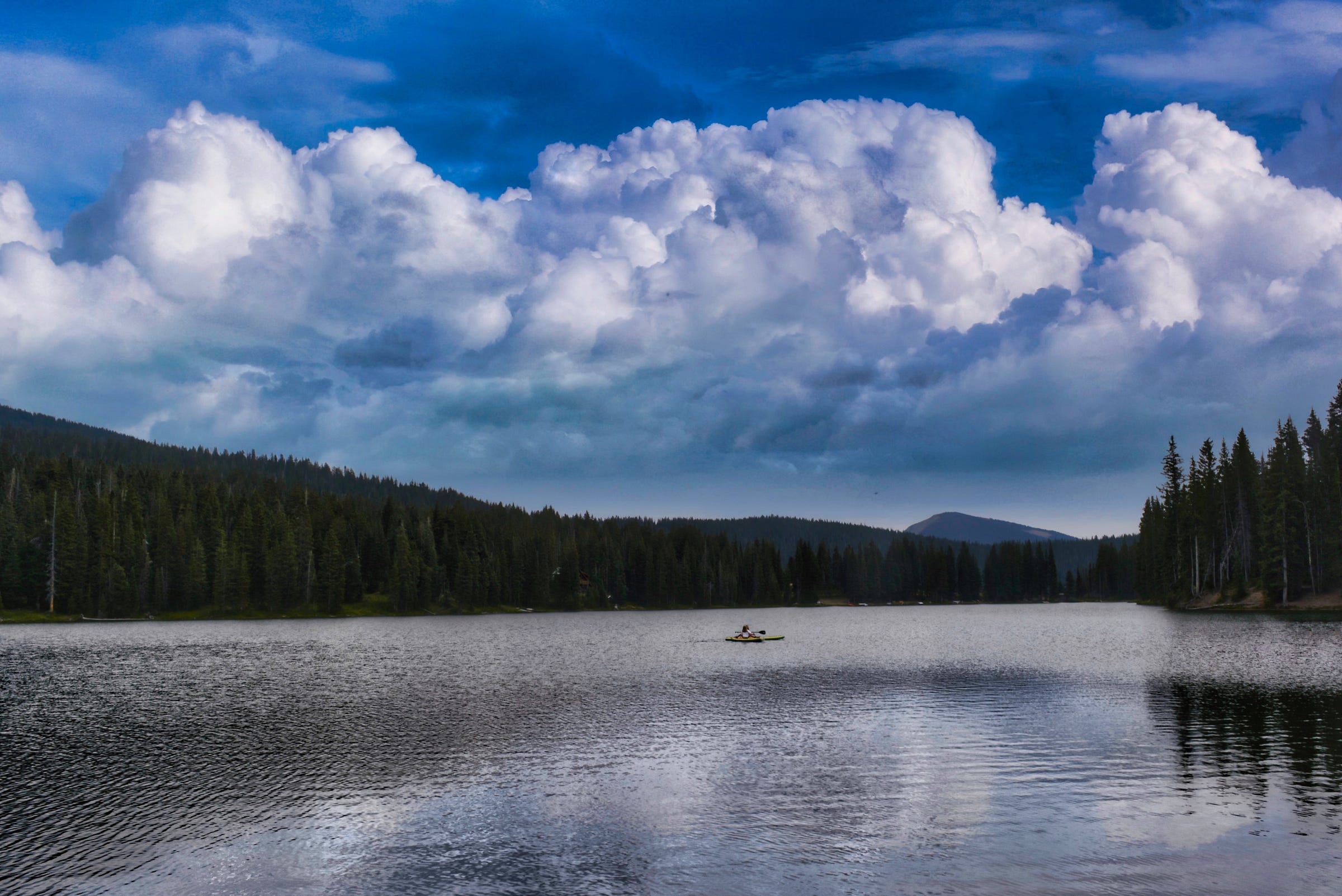 A quiet mountain lake with rolling white clouds against a darkening sky; a small kayak in the middle of the lake