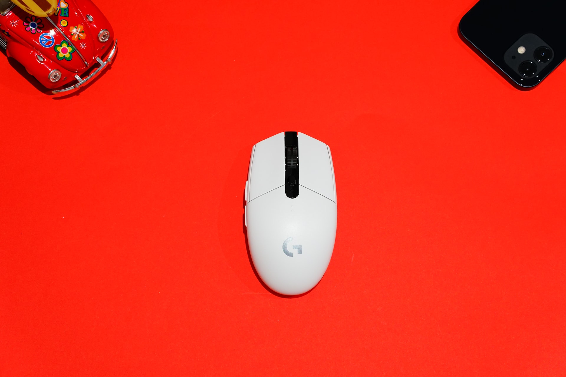Un mouse bianco con il logo di Google al centro, su sfondo rosso. Dagli angoli superiori dell'immagine spuntano una macchina giocattolo rossa con decorazioni floreali e un cellulare con la cover nera.