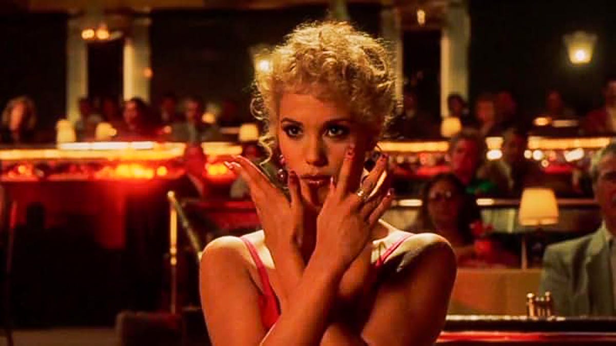 Elizabeth Berkley as Nomi Malone stands and strikes her iconic inverted jazz hands pose while watching Goddess at the Stardust for the first time and imitating the dance moves seen on stage. She's framed by neon reds and soft lamp light with the rest of the audience seated behind her. Elizabeth Berkley as Nomi Malone stands and strikes her iconic inverted jazz hands pose while watching Goddess at the Stardust for the first time and imitating the dance moves seen on stage. She's framed by neon reds and soft lamp light with the rest of the audience seated behind her.