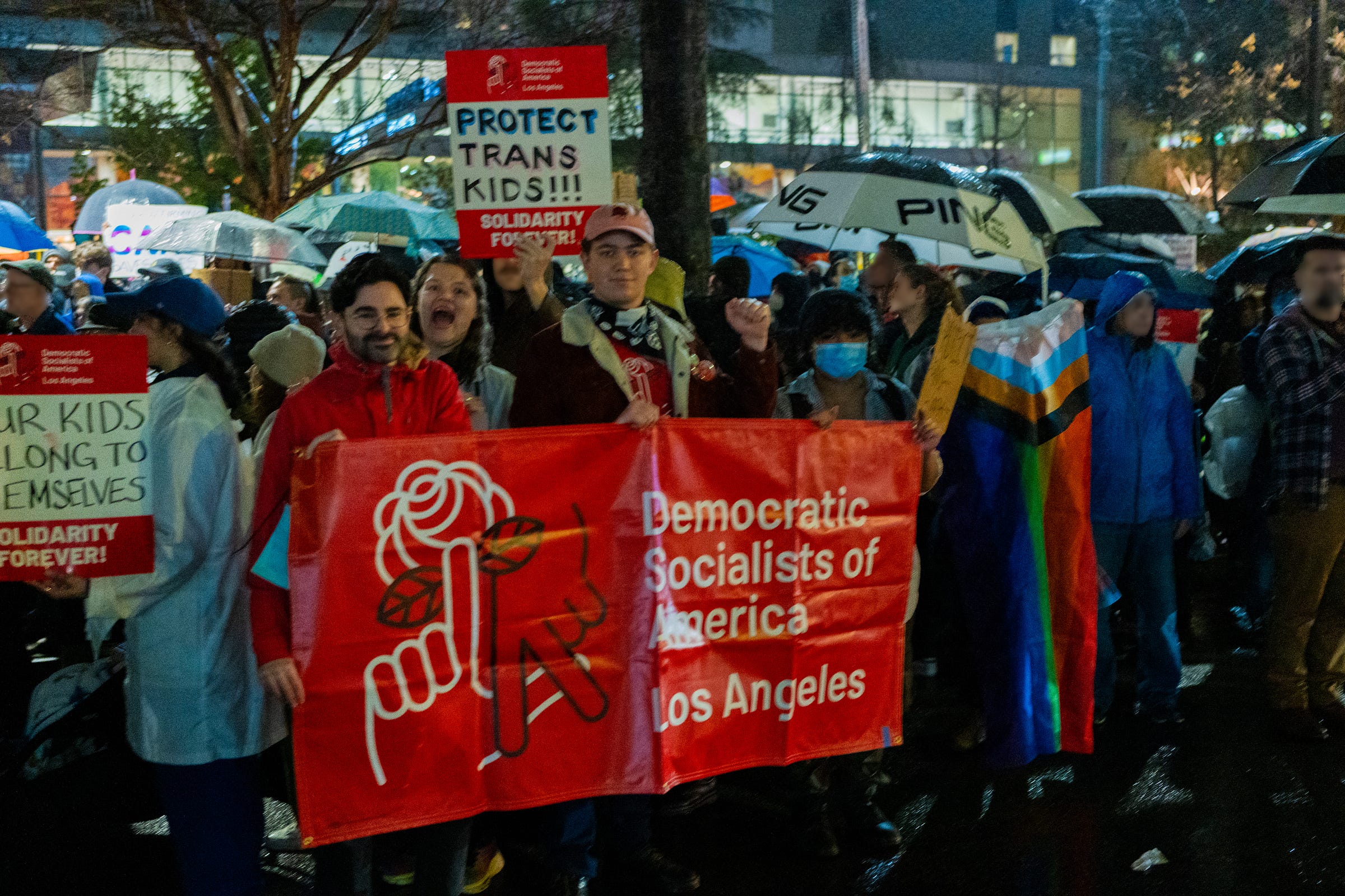 Four DSA members hold a red Democratic Socialists of America Los Angeles banner in front of a crowd of protestors. Several signs read "Solidarity forever!" and "Protect trans kids!"