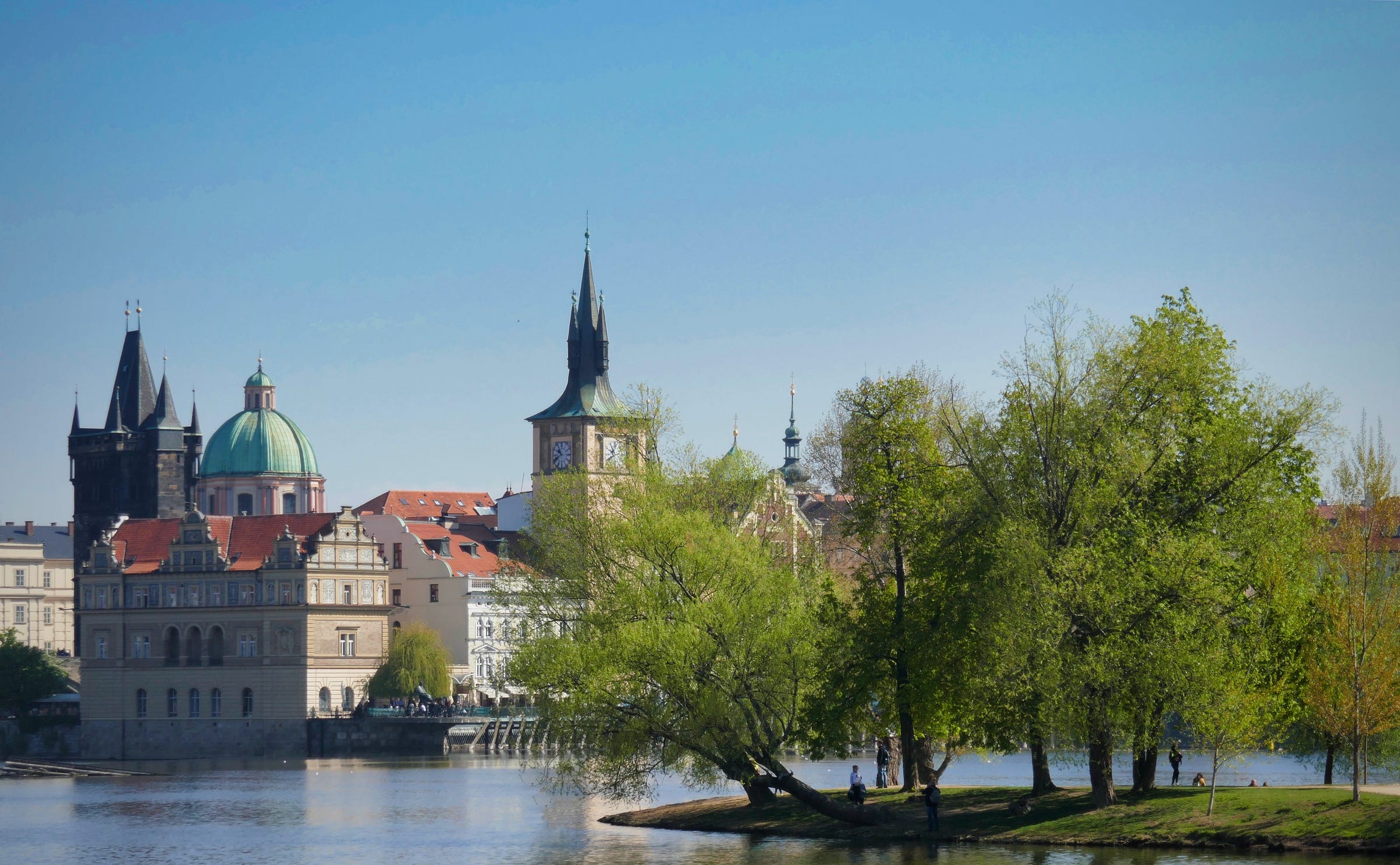 A view of a small island with trees and a park bench close to the shore of the Vltava River in Prague. In the background are buildings including a clock tower.