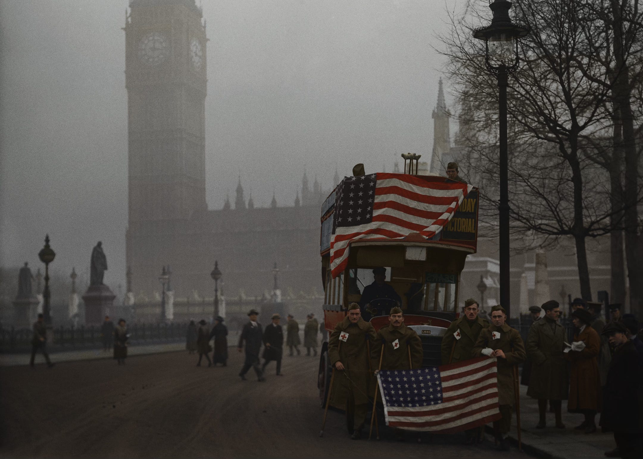 Colorized photograph from 1918 shows a group of wounded American soldiers posing on Parliament Square in London on an overcast November day. An American flag is draped over the red bus behind them. Big Ben and the Houses of Parliament in the background.