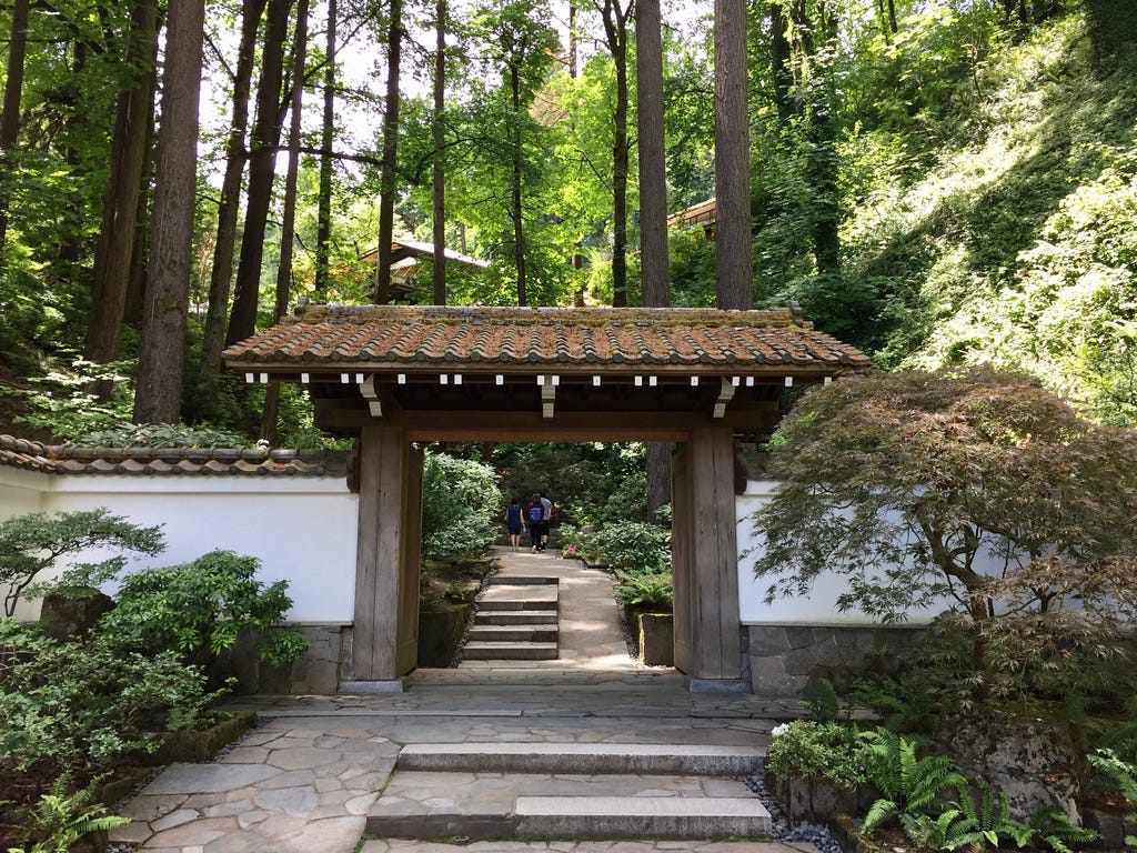 A Japanese garden gate with tiled roof and white stucco wall, stone path starts and contues through it, a forested hillside beyond beckons with lush green vegetation.