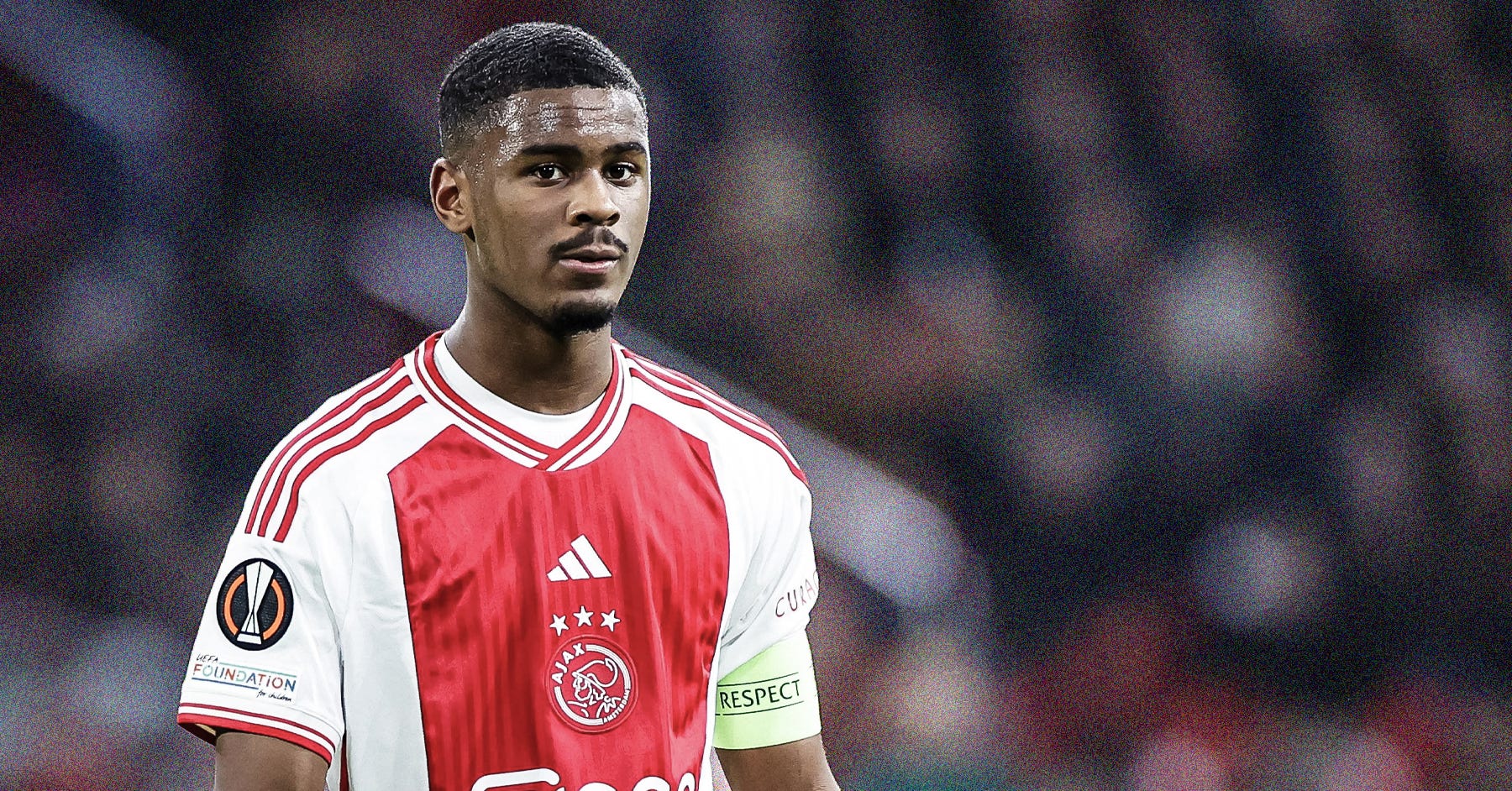 A photo of Jorrel Hato wearing a red-and-white AFC Ajax shirt and a yellow captain's armband. He's looking toward the camera with a pensive look on his face. Behind him are green and white circles, the latter with the SCOUTED50 logo inside it.