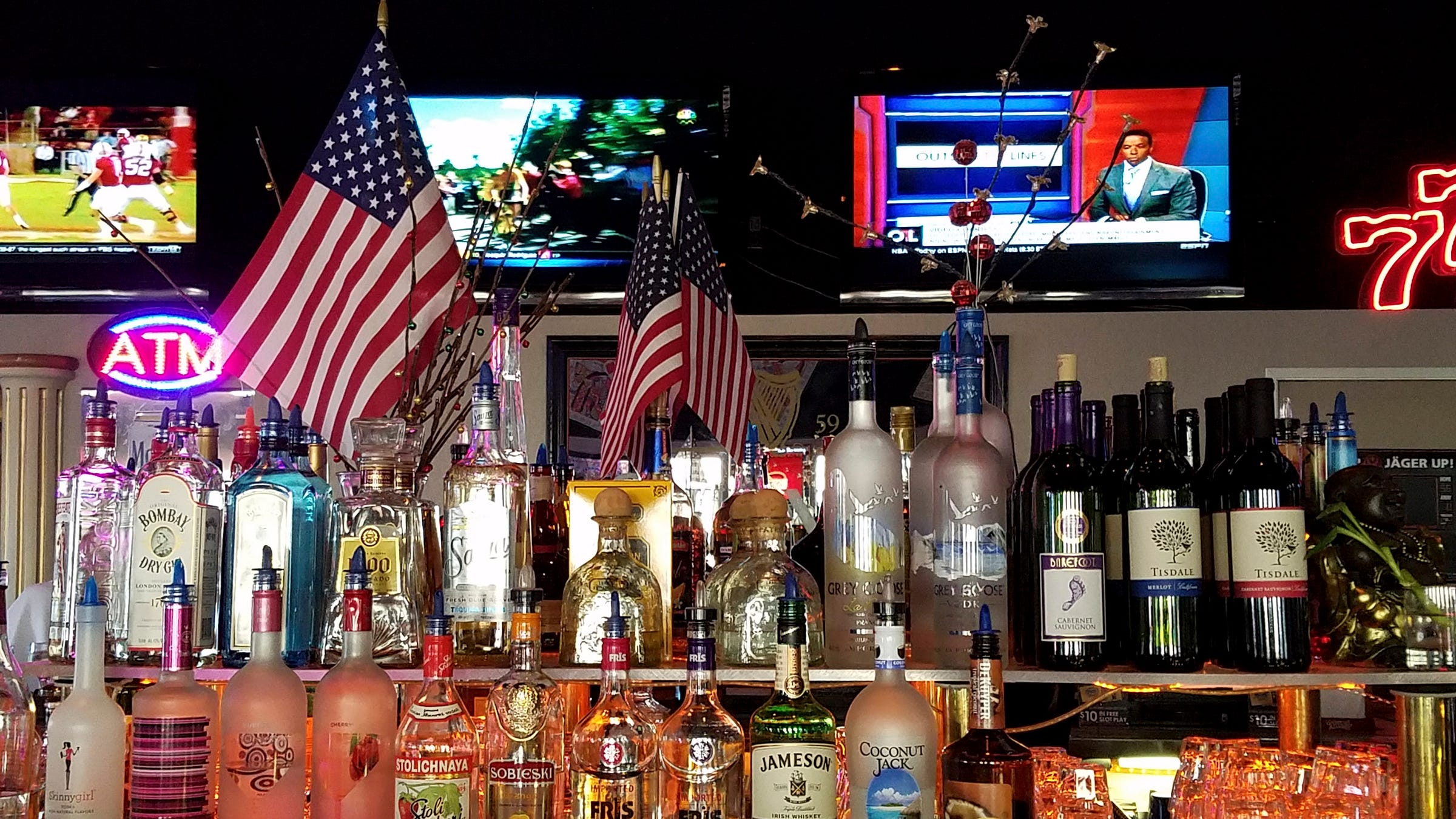 Photo of a bar. In the foreground are two stacked rows of bottles, including some corked bottles of wine and mostly liquor bottles with spouts for easy pouring. Behind  the bottles are a handful of US flags. Behind them, against a distant wall,  is a gaudy white architectural column with gold fluting and a marble capital. The column is simple stuck to the wall and is not supporting anything. Beside it is a neon sign, "ATM" in red letters sitting inside a blue neon oval. Another neon sign "77" sits to the right. At the top of the wall are three large, flatscreen televisions showing a football game, a bicycle race, and a sports reporter talking to camera in a studio setting.