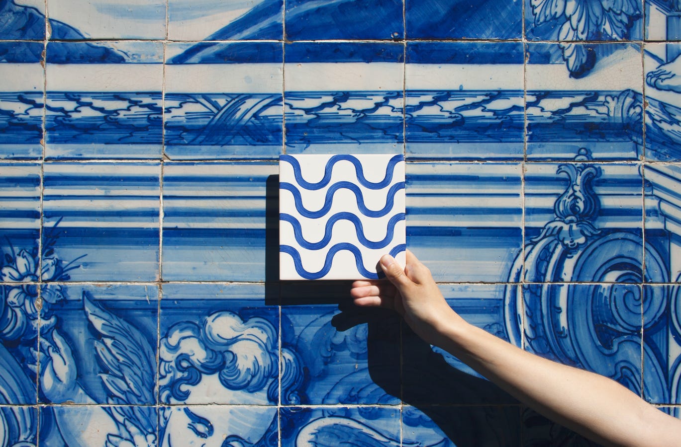 Uno scorcio di una parete di Porto, decorata con le tradizionali piastrelle bianche e azzurre. Al centro dell'immagine, una mano regge una piastrella bianca decorata con onde azzurre.