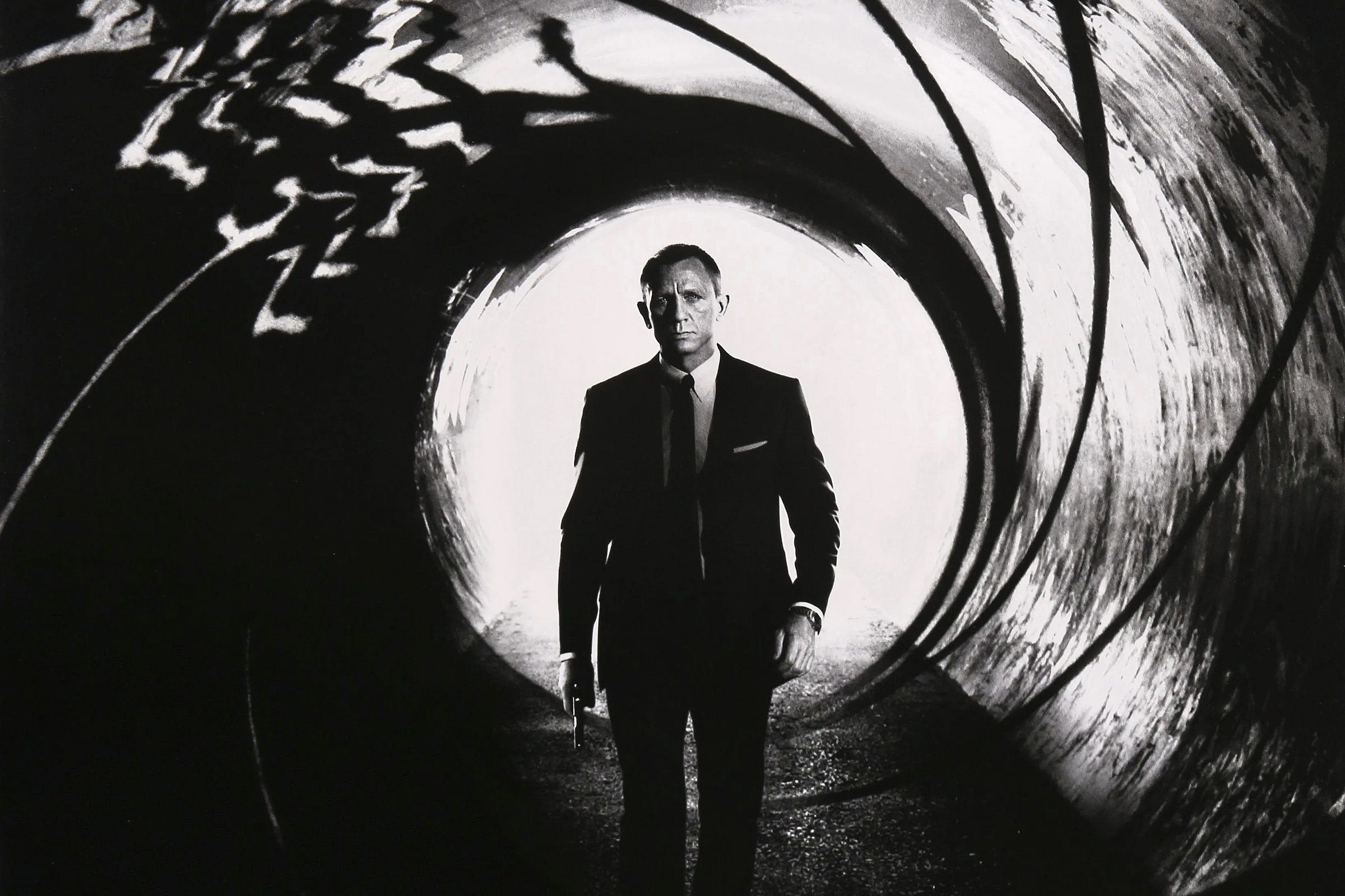 Black and white photograph of the actor Daniel Craig as James Bond walking through a circular tunnel carrying a pistol