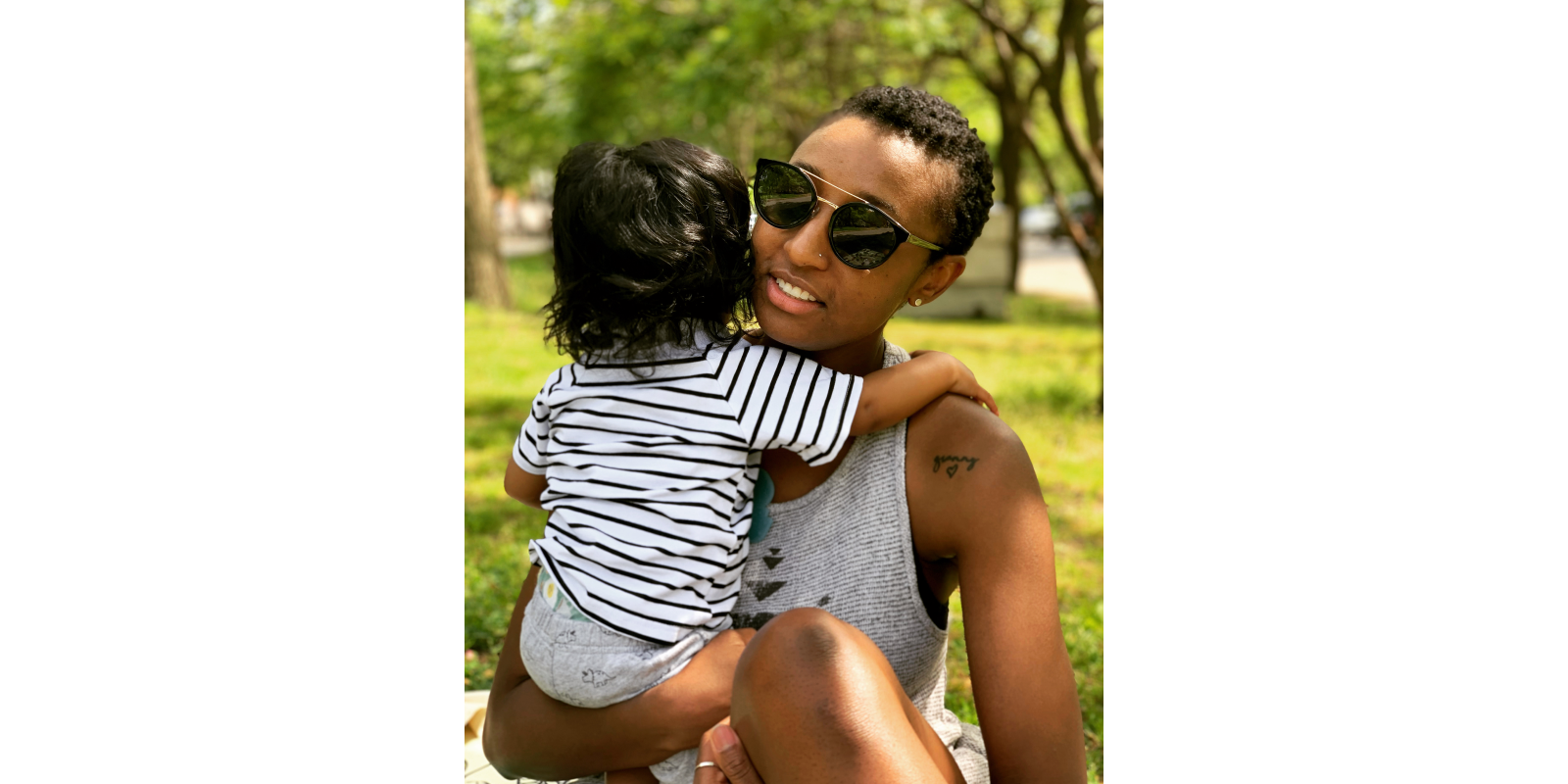 Portrait of Ashley Woodfolk in sunglasses and tank top holders her son who is facing the other direction in a black and white striped t-shirt.