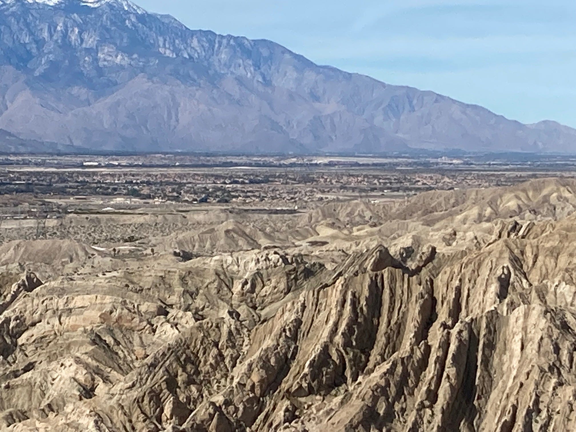 Rugged badlands and striated cliffs in the foreground near Coachella give way to desert communities in the midground and towering mountains in the background.