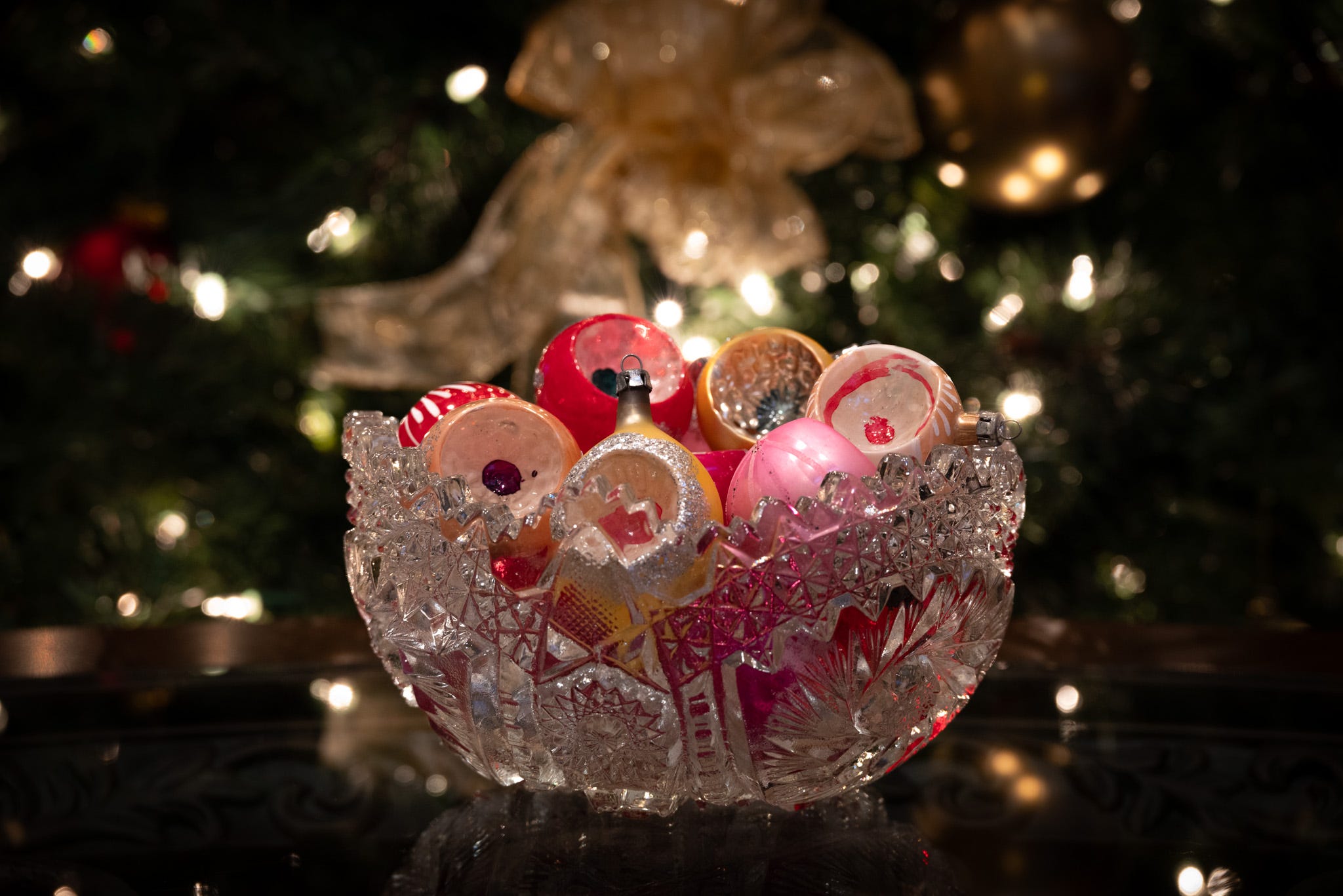 A crystal bowl full of antique glass ornaments with the lights and bows of a christmas tree in the background