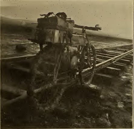 Old black and white photo of George A. Wyman's California motorcycle on railroad tracks. The bike and the ground are extremely muddy. The motorcycle is resting on adjacent railroad ties and leaning against one of the iron rails. The motorcycle has a rear rack upon which a pair of rectangular cases are strapped. The is no ballas or gravel between the railway ties.