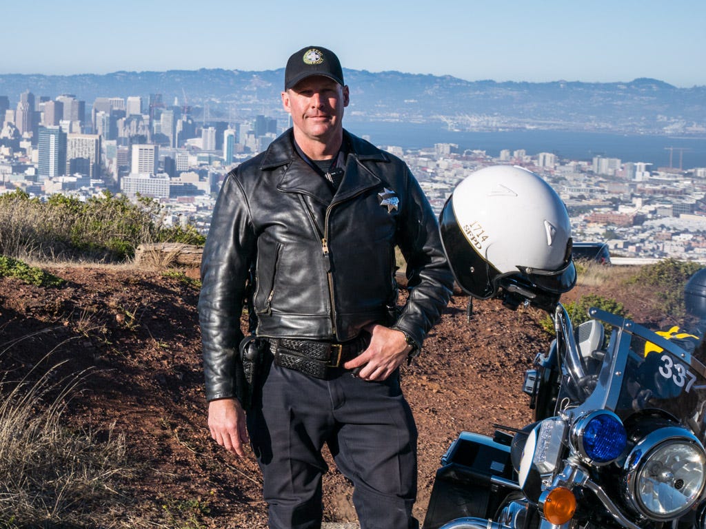 A San Francisco Police Department officer poses beside his Harley-Davidson police cruiser on Twin Peak in San Francisco. Officer Gene Cornryn is wearing a motorcycle police officer's uniform consisting of dark blue slacks, a black leather braided belt and gun holster, a black leather biker's jacket with brass zippers and an officer's badge on the left chest, and a black baseball cap with the SFPD logo. Officer Cornryn's left thumb is tucked into his belt as he looks directly at the camera with a closed-mouth smile. To his left, his Harley-Davidson motorcycle faces the camera. The headlight and windscreen is visible in the lower right corner of the frame, with the right round amber turn signal and larger round blue flashing light above it. Officer Cornryn's white and black three-quarter motorcycle helmet sits casually on the right handlebar of the motorcycle. Behind him is a patch of dirt and some short scrubby foliage, and in the hazy distance are the towers of downtown San Francisco, San Francisco Bay, Oakland, and the foothills beyond.