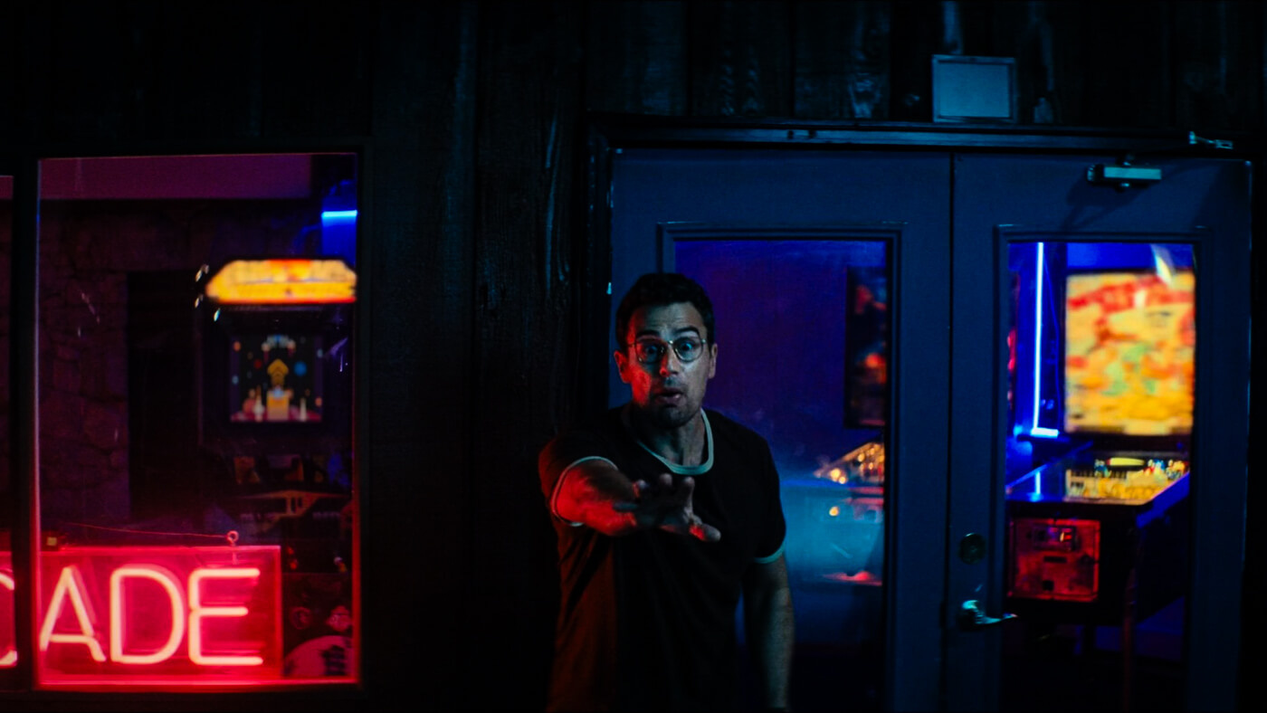 Theo James, wide-eyed and mid-gesture, reaches forward to quickly warn someone outside a neon-lit arcade with vintage video game machines.