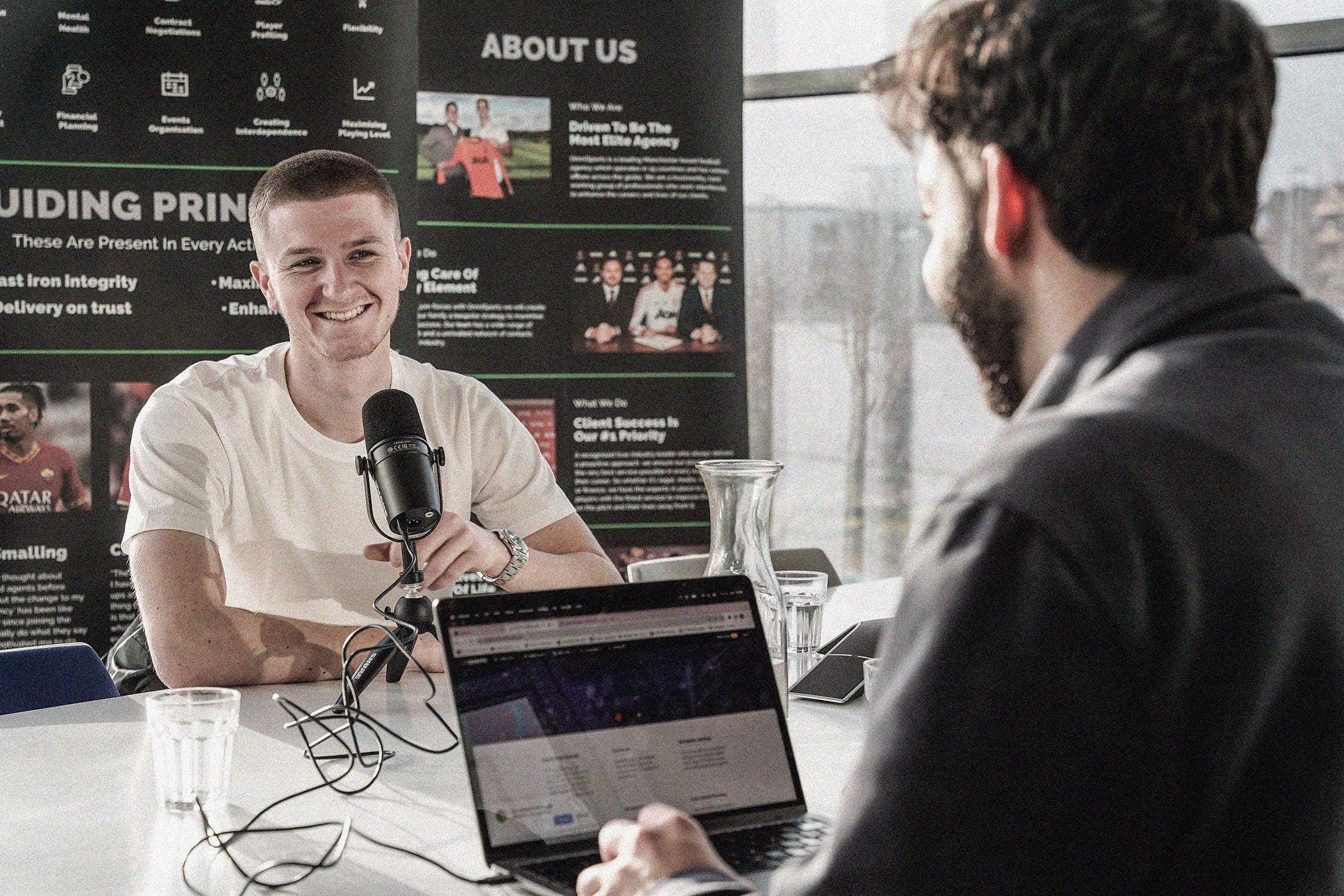 A photo of a smiling Adam Wharton sat at a white table in front of a microphone. In the foreground is Phil Costa, SCOUTED journalist, sat at a laptop.
