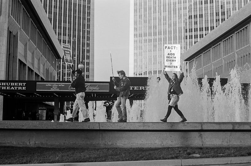 Three activists walk along a fountain outside Century City Mall, one of Los Angeles’s most iconic shopping and movie centers. They hold signs saying "FACT: 1 AIDS DEATH EVERY 7 MINUTES" and other illegible signs. The backdrop is of a fountain, tall buildings, and a movie theater.