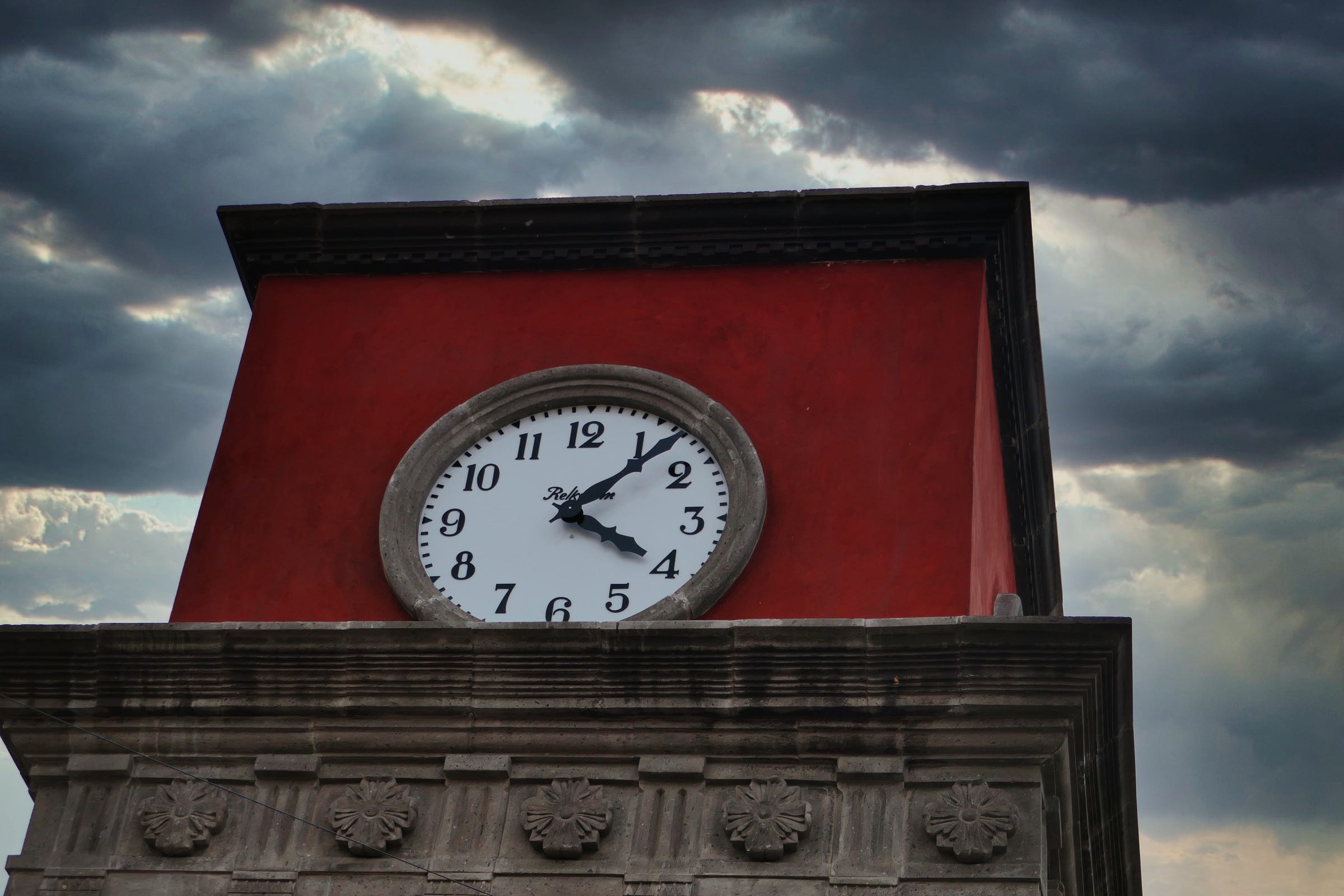 A clock frozen at 4:07 p.m on a red steeple with dark stormy clouds behind it