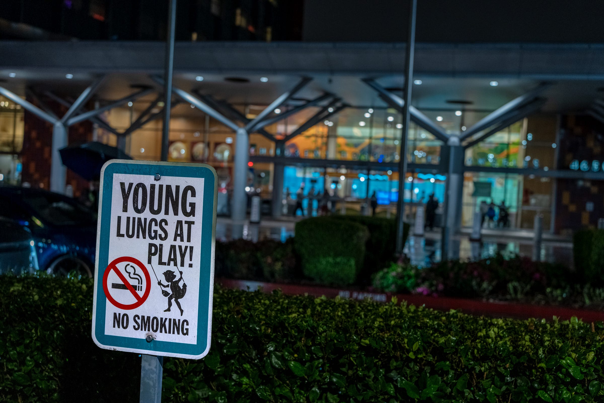 A sign reads "YOUNG LUNGS AT PLAY! NO SMOKING" in front of a row of hedges by the hospital entrance