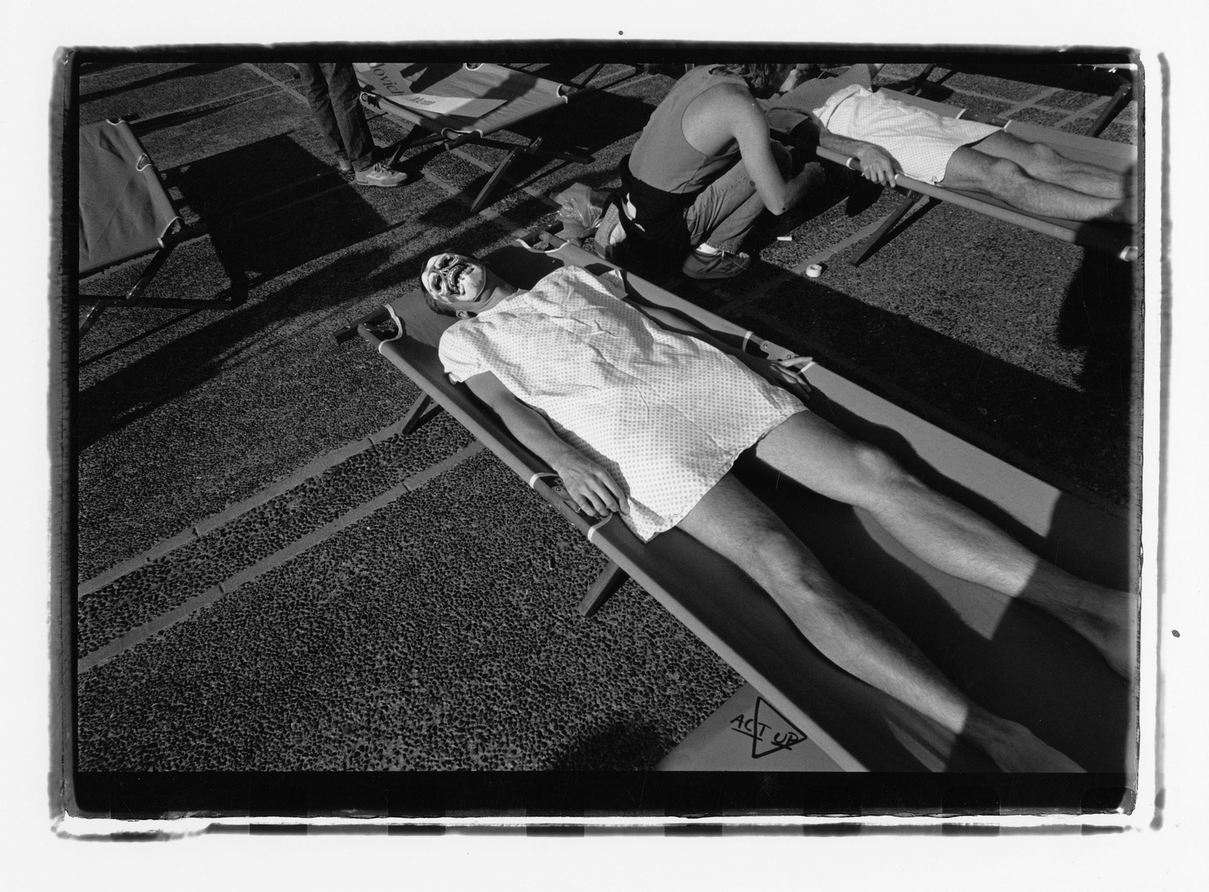 A person wearing a skull mask and hospital gown lies on a stretcher surrounded by several other stretchers in a parking lot. A hand drawn triangle with the words “ACT UP” is visible by the foot of the front stretcher.