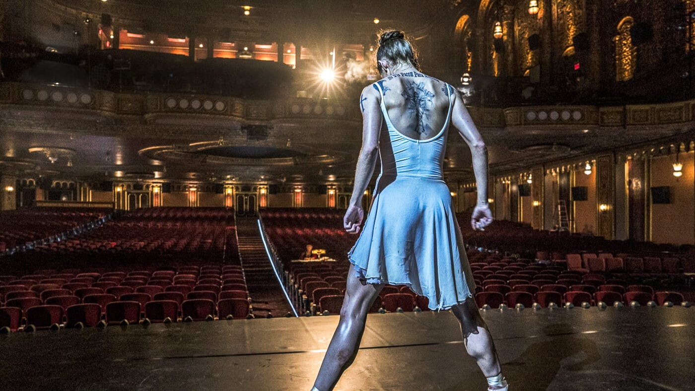A female ballerina shown from the rear in a white leotard stands alone onstage, tattooed muscular back exposed, facing an empty velvet-seated theater.