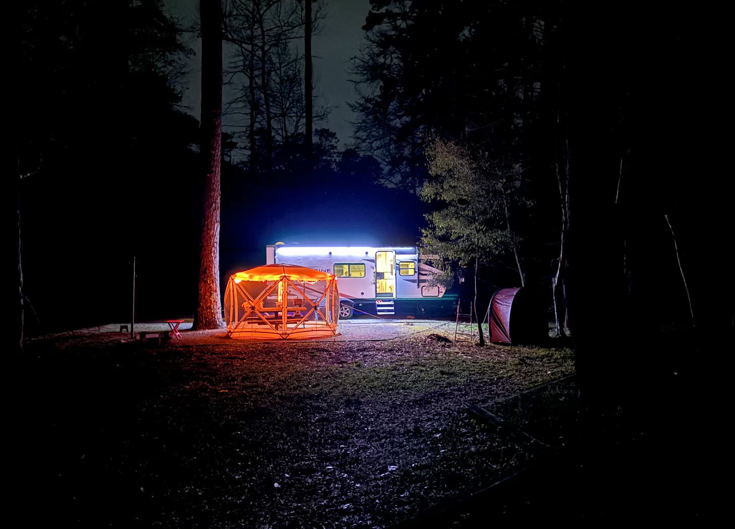 travel trailer and tent brightly lit surrounded by dark woods at night