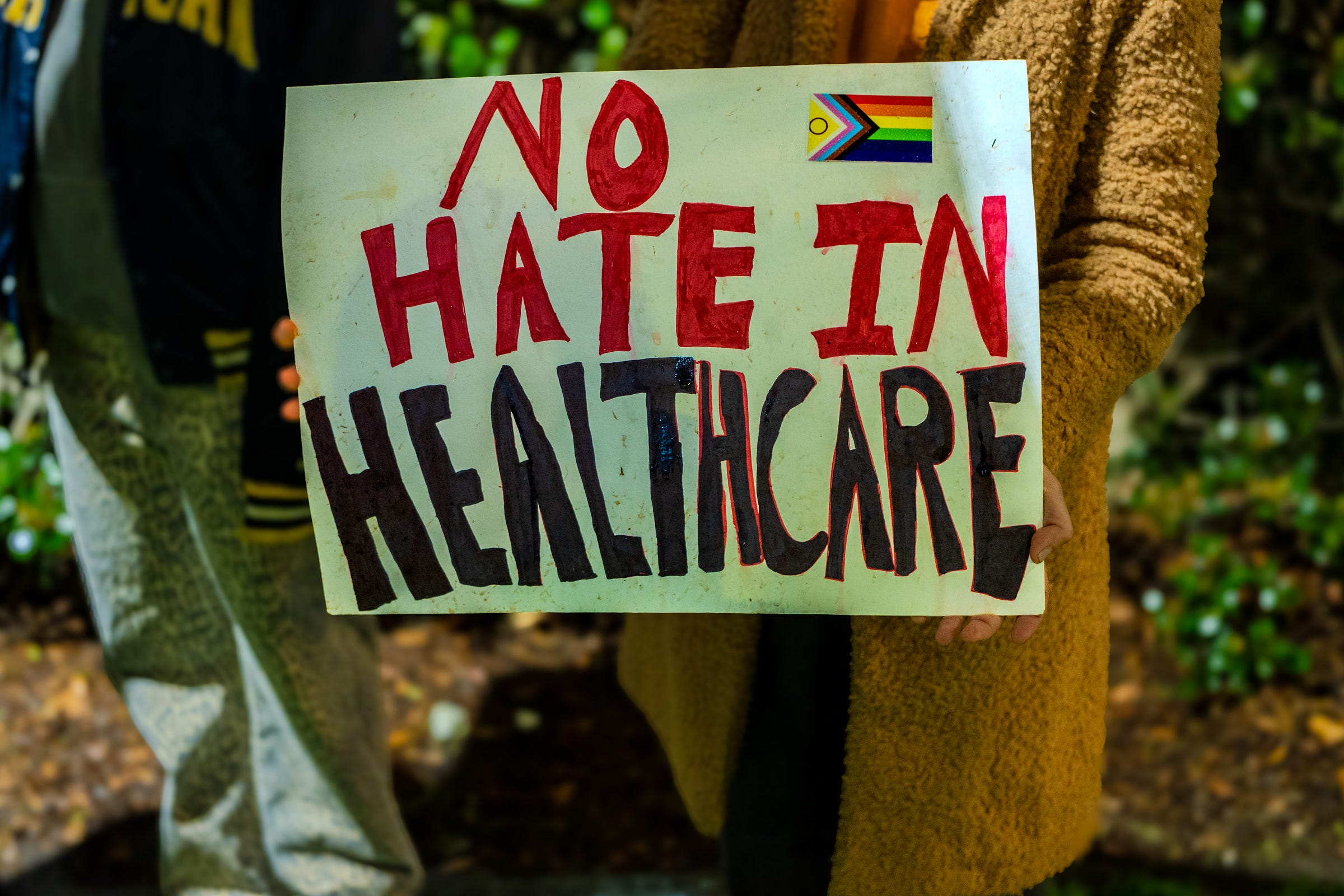 A sign with an intersex-inclusive Pride Progress flag sticker reads "NO HATE IN HEALTHCARE"