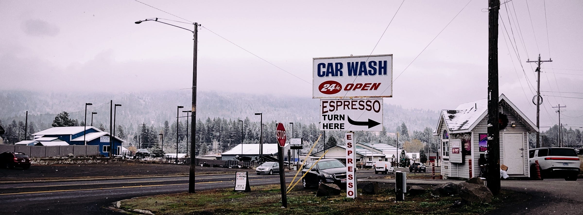 Photograph of a road in North Idaho. In the foreground is a sign for a car wash, advertising espresso. In the background are small buildings and trees dusted in a light coating of snow.