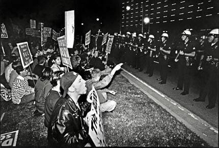  A black and white photo of a crowd of ACT UP activists kneeling on the grass holding protest signage as they face off with a line of police officers wearing helmets and holding weapons.