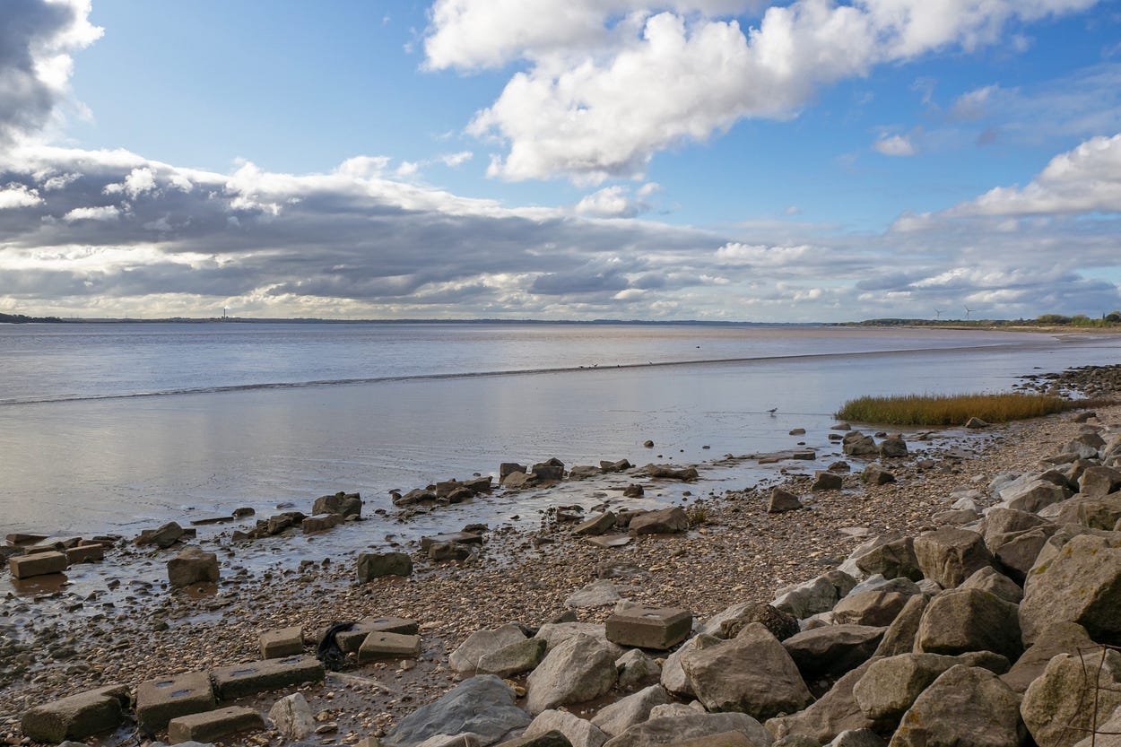 Photograph of the foreshore of the Humber Estuary. There are boulders in the foreground, and the tide is a little way out. There is a blue sky with clouds abovel