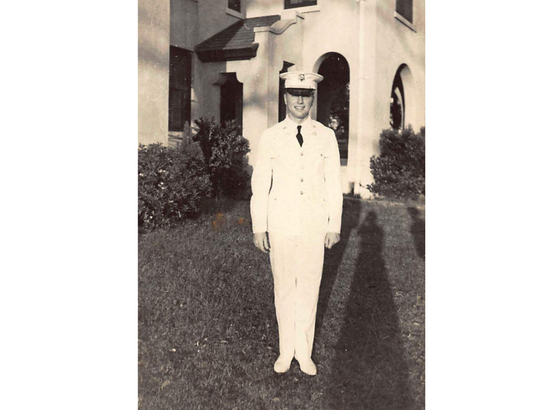 A young naval officer in dress uniform standing in front of a building with a shadow of the photographer visible to on the right.