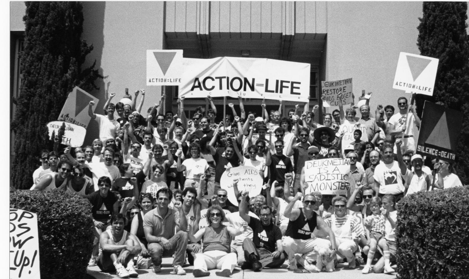 A black and white group photo of more than a hundred ACT UP activists raising their fists and protest signage at the entrance of a building. Various signs read “Action = Life,” “Silence = Death,” “Deukmejian restore AIDS Budget Cuts,” and “Deukmejian is a sadistic monster.”