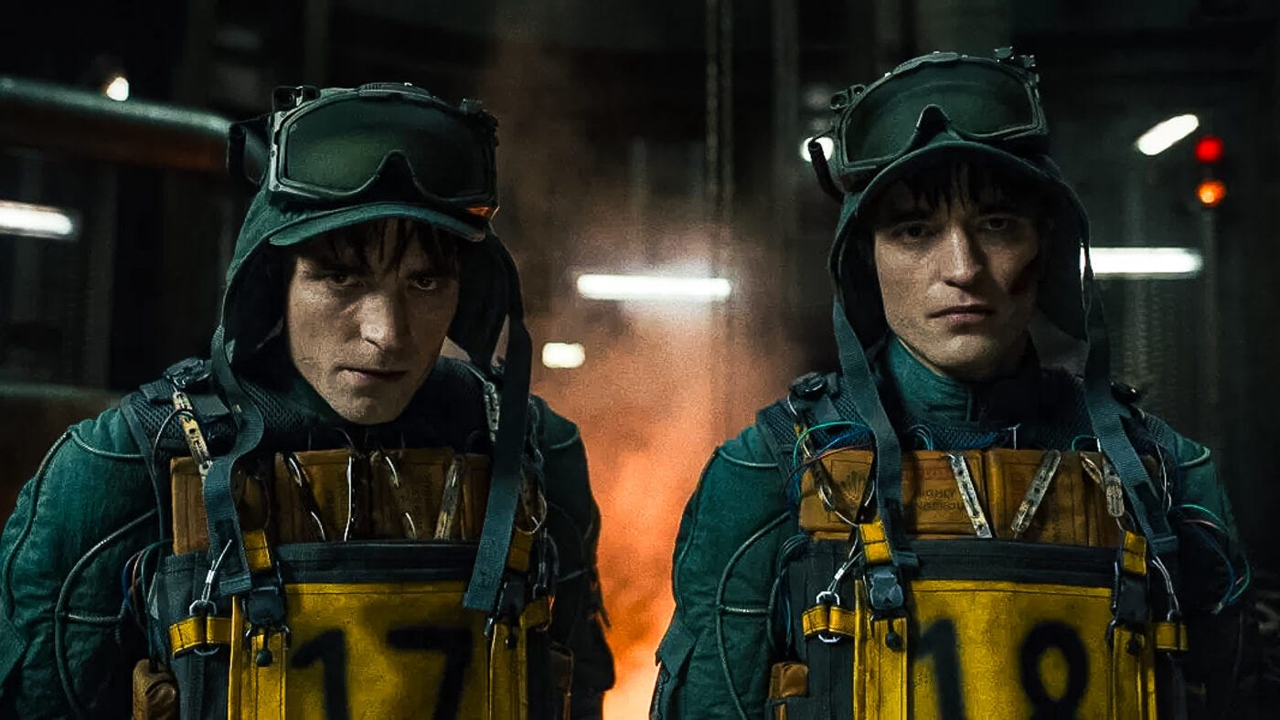 Two versions of Robert Pattinson stand in front of an incinerator, wearing numbered vests marked “17” and “18,” suited in green survival gear with goggles and harnesses.