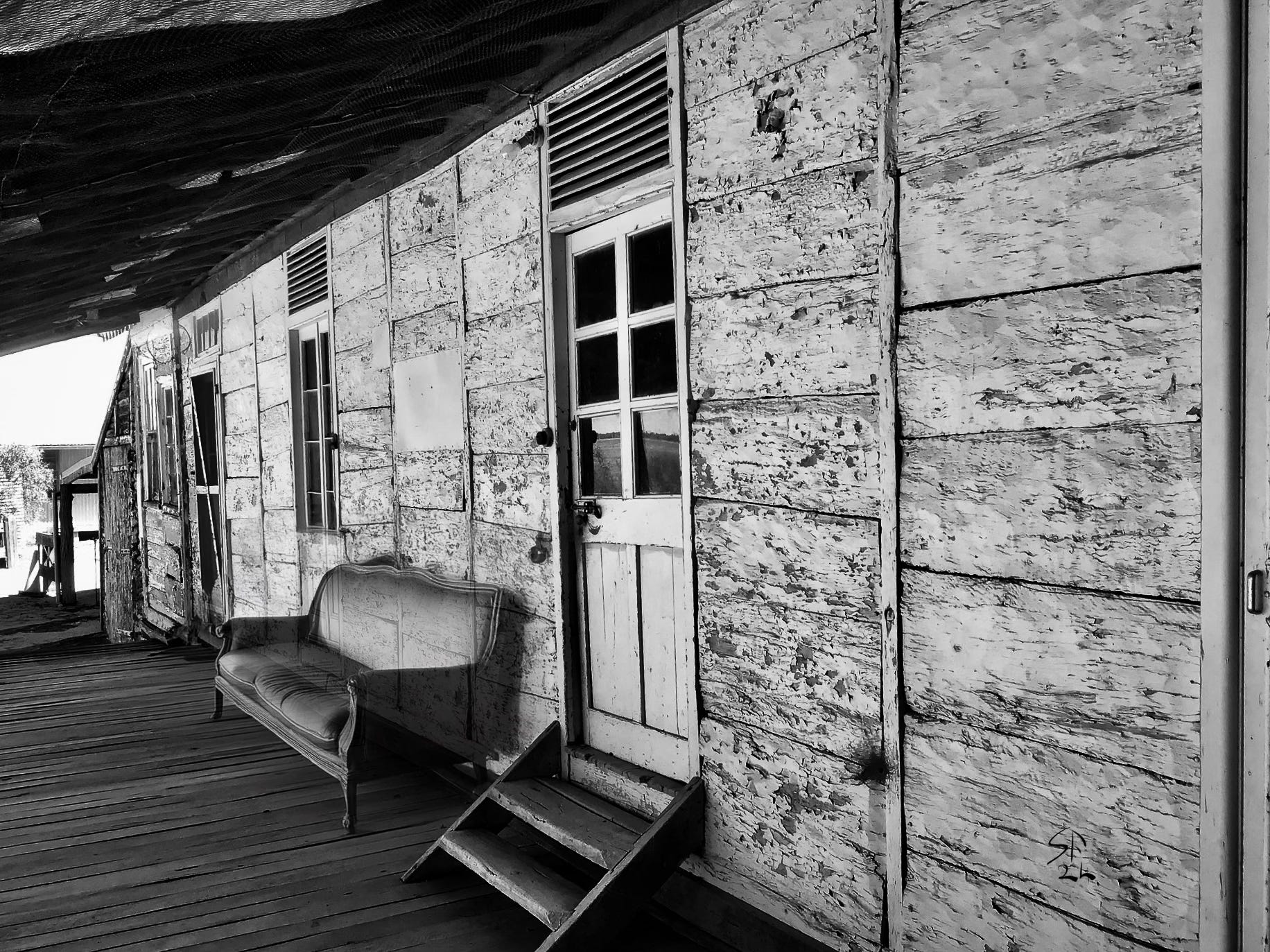 Black-and-white photo: old Australian farmstead veranda; next to the door is a shabby old sofa. The wall of the house is visible through the sofa. Is it there or not?