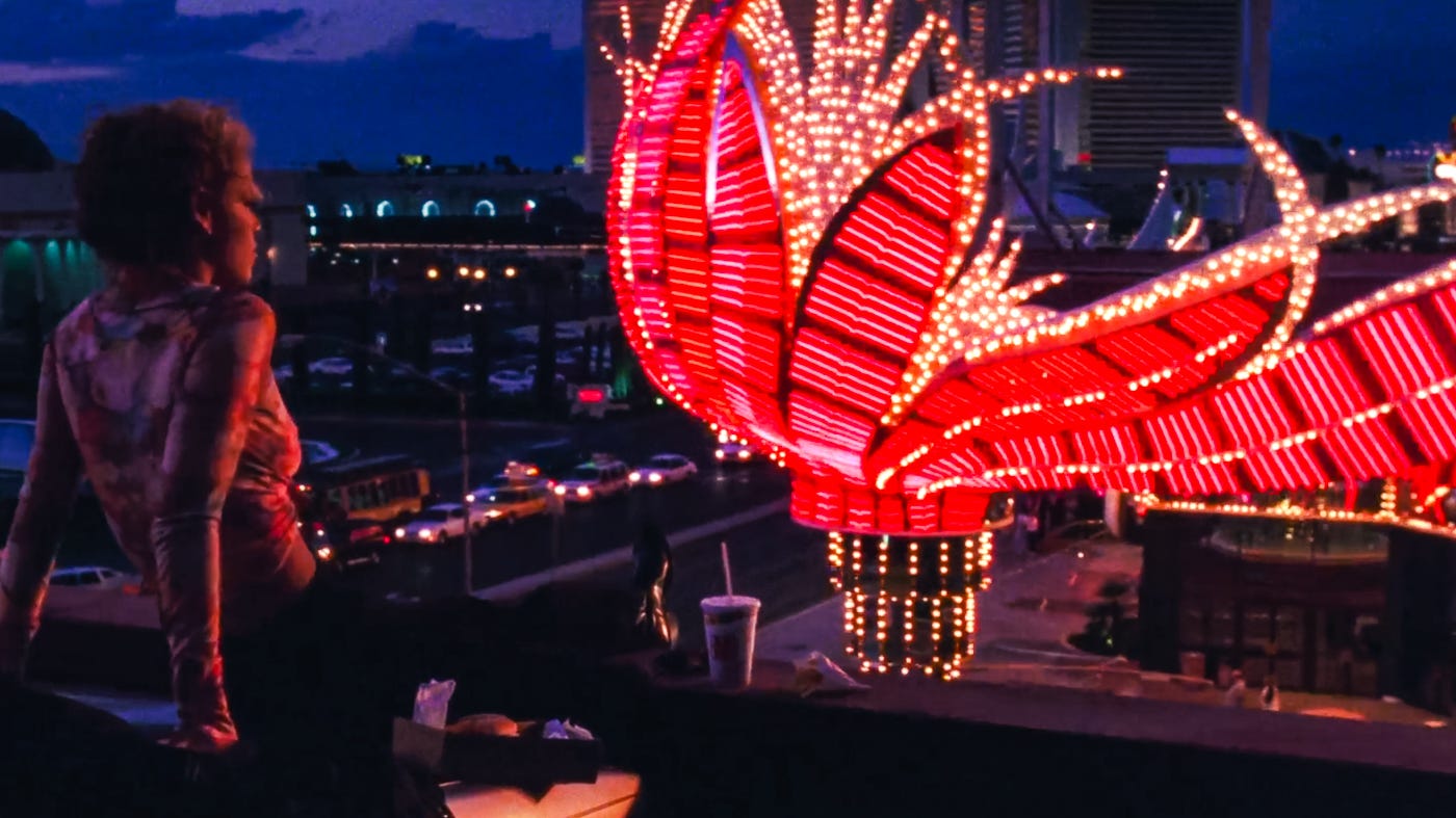 Nomi Malone sits on a rooftop in a sheer top, silhouetted against the glowing pink and gold lights of the Flamingo marquee on the Las Vegas Strip at dusk.