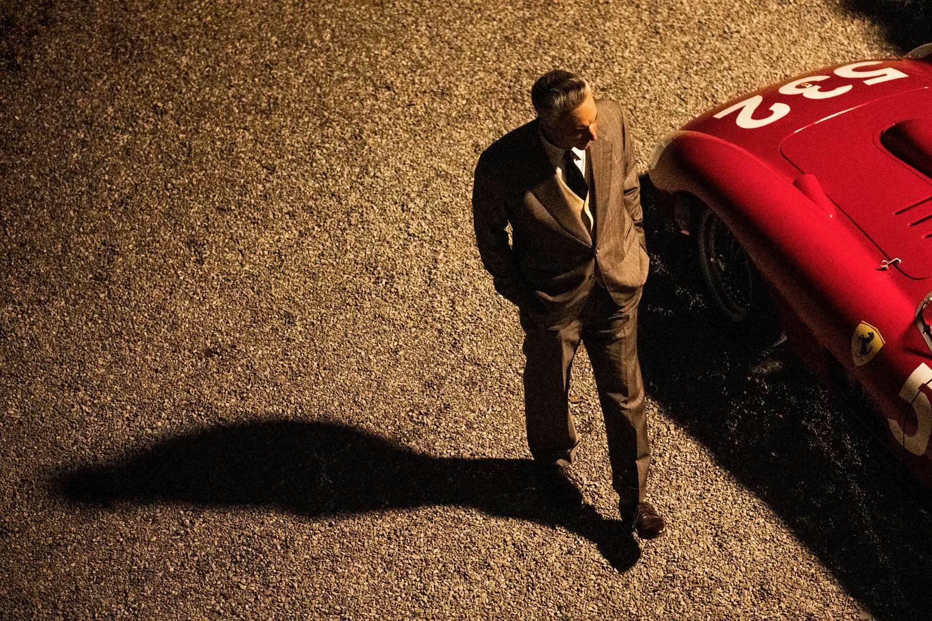 An overhead shot of Enzo Ferrari (Adam Driver) walking past a racecar