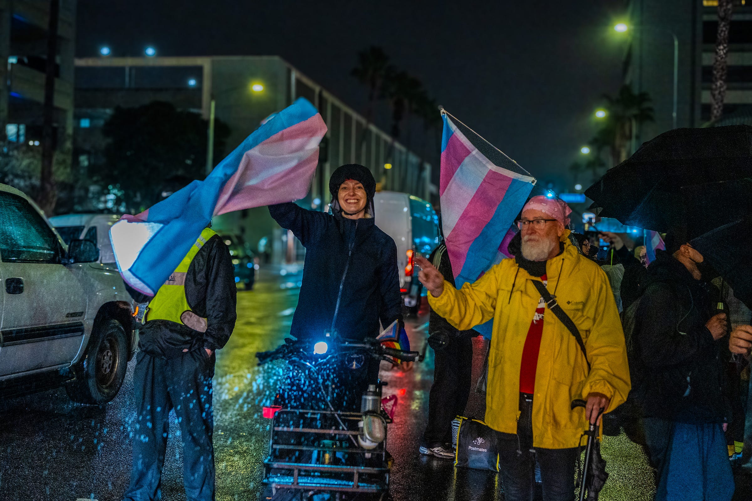 Several protestors on the street waving trans flags and protesting. One protestor is on a bike.